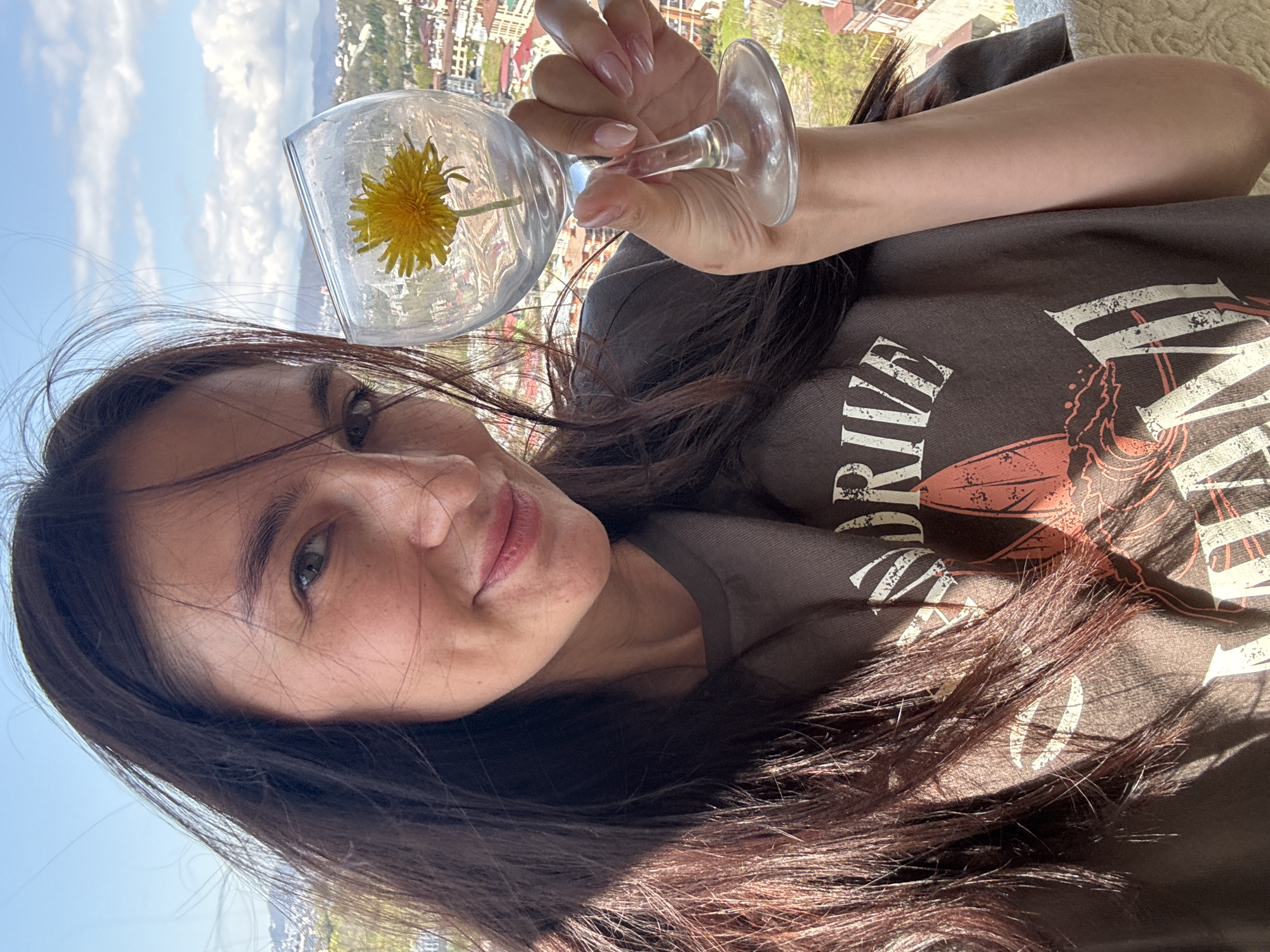The image shows a woman holding a wine glass with a single dandelion flower inside. She appears to be taking a selfie, as the camera is positioned close to her face. She has long, dark hair and is wearing a brown t-shirt with a faded graphic. The background reveals a cityscape with a clear, blue sky and distant mountains. Based on the style of buildings and surrounding geography, the location is consistent with Sochi, Russia.