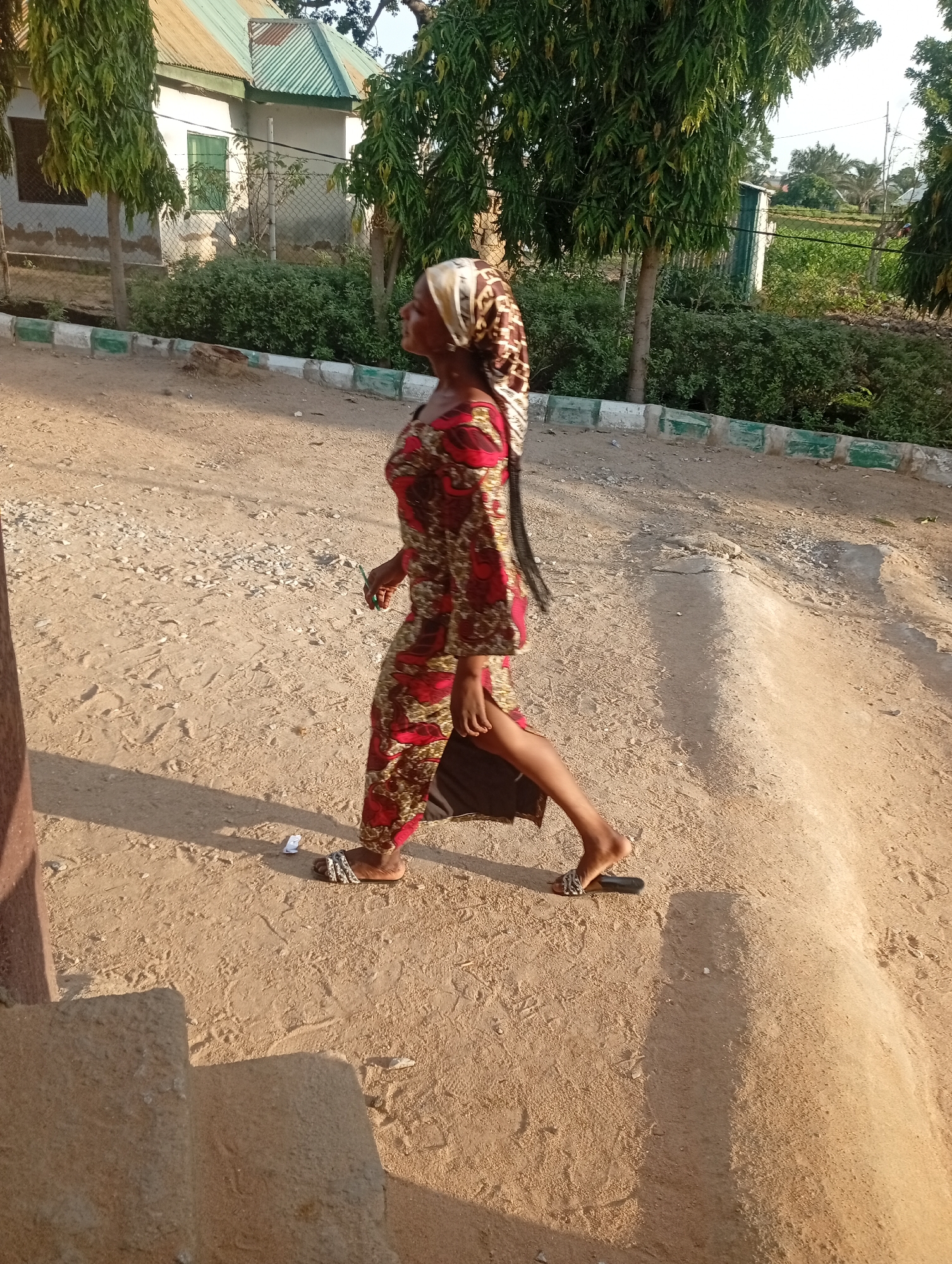 A person wearing a red and gold patterned dress and a headscarf walks on a dirt path. The person is walking from left to right, in profile. They are wearing sandals. In the background, there is a building with a green, corrugated metal roof and some trees. The ground is dirt and gravel. The scene appears to be outdoors during the day, with shadows suggesting it is late afternoon. There is no visible text.