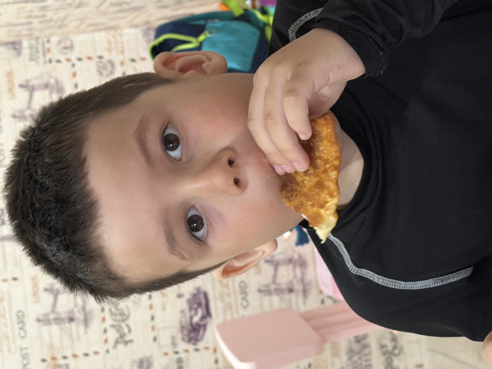 A child is eating food, possibly an egg roll or similar fried pastry. The child has brown hair and is wearing a black, long-sleeved shirt with light gray stitching around the collar. The background shows a printed tablecloth with various images, including what appears to be a postcard theme. Behind the child's head is a colorful backpack. A pink chair or object is partially visible near the bottom of the frame. The photo was taken indoors. The location in the city of Chaikovsky, Russia is undetermined.