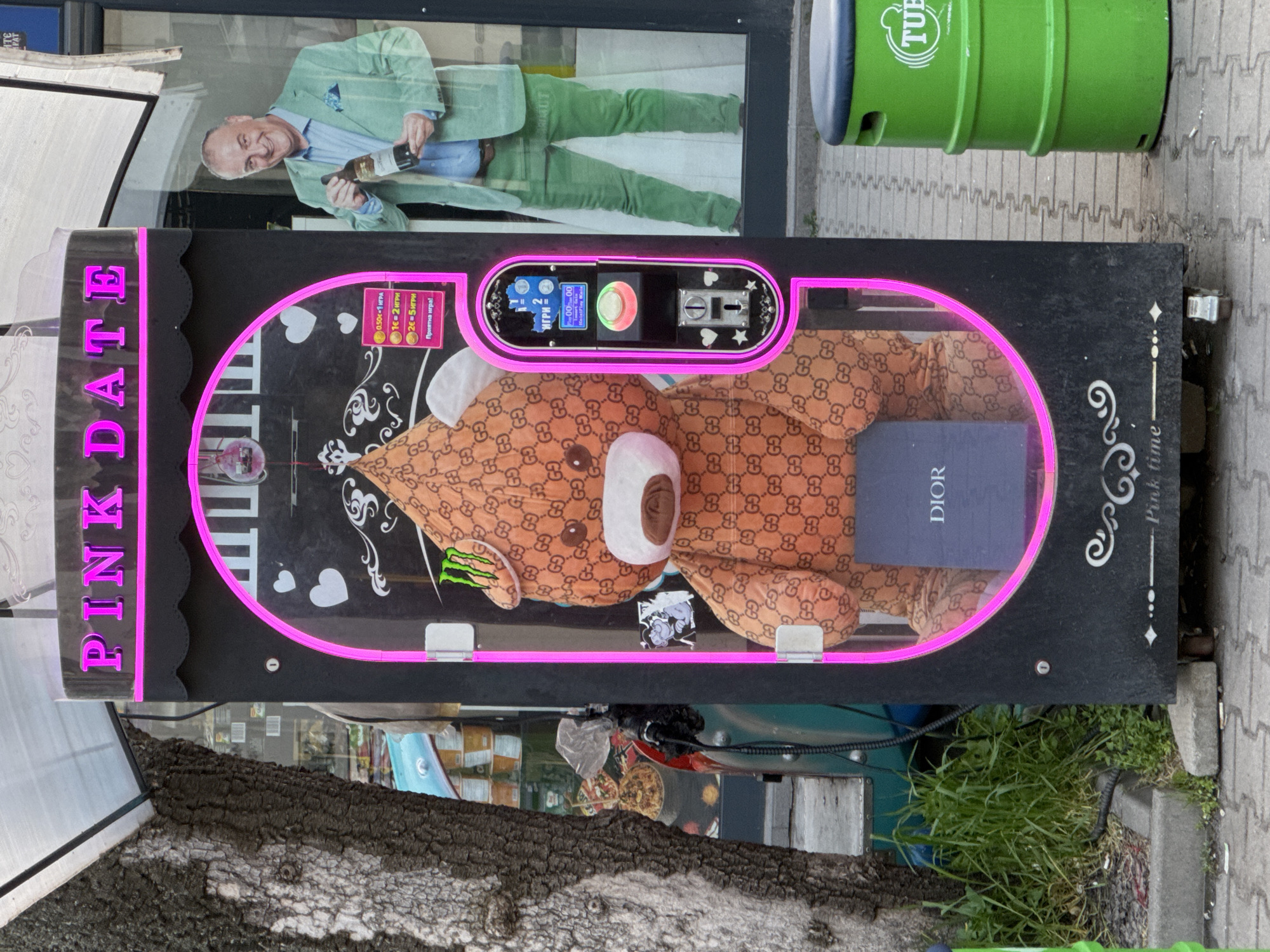 The image shows a "Pink Date" claw machine on a sidewalk in Sofia, Bulgaria. Inside the machine, a Gucci-patterned teddy bear lies next to a Dior box. The machine has a pink neon outline. The control panel indicates the game costs 0.50€ for one play, 1€ for two plays, and 2€ for five plays. A green "TUB" recycling bin is visible in the background. A storefront is reflected in the glass, showing a man in a green suit holding a bottle. Trees and sidewalk details are also visible in the shot.