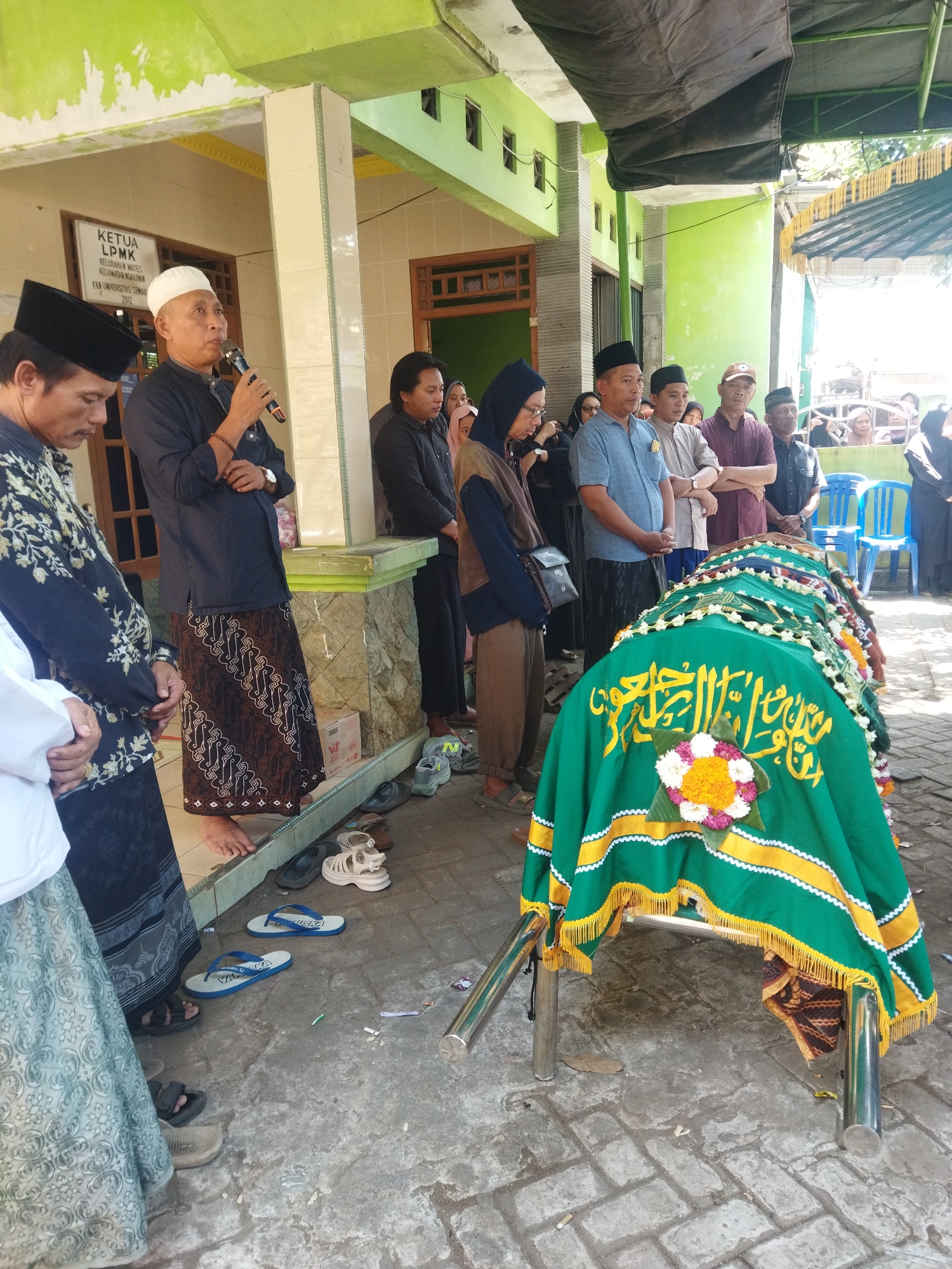 In Semarang, Indonesia, an outdoor scene depicts a funeral service. A coffin, draped in a green cloth adorned with yellow trim and Islamic calligraphy, rests on a metal stand. Several men, some wearing traditional Indonesian attire like sarongs and peci hats, stand in a semi-circle, some observing the coffin and others listening to a man speaking into a microphone. Behind them, more people are visible, some sitting on chairs. The background shows a building with green walls and a covered walkway. The ground is paved with interlocking bricks and scattered with various footwear, including sandals and flip-flops. The atmosphere appears somber and respectful.