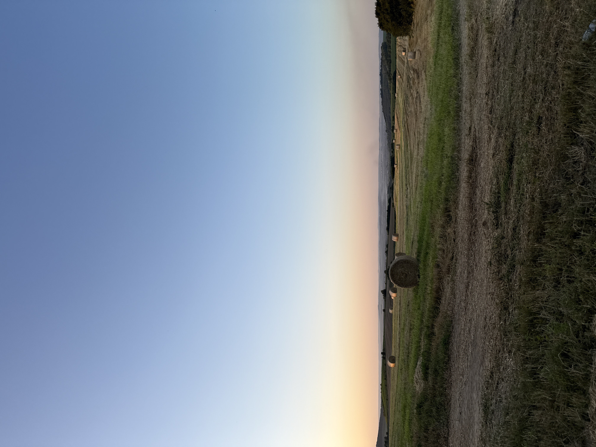 A wide shot captures a rural landscape at dusk. The sky is a gradient of soft blue transitioning to a pale orange at the horizon, with no discernible celestial bodies visible. In the foreground, a field is covered with cut grass and straw, with some taller, dry grass present. Further into the midground, a large field features numerous cylindrical hay bales scattered across its surface. The field itself is a mix of green grass and mown areas. In the distance, rolling hills are visible, dotted with trees and vegetation. The overall lighting suggests the late evening hours. There are no people or animals visible in the scene. The location is identified as Unknown, Unknown.