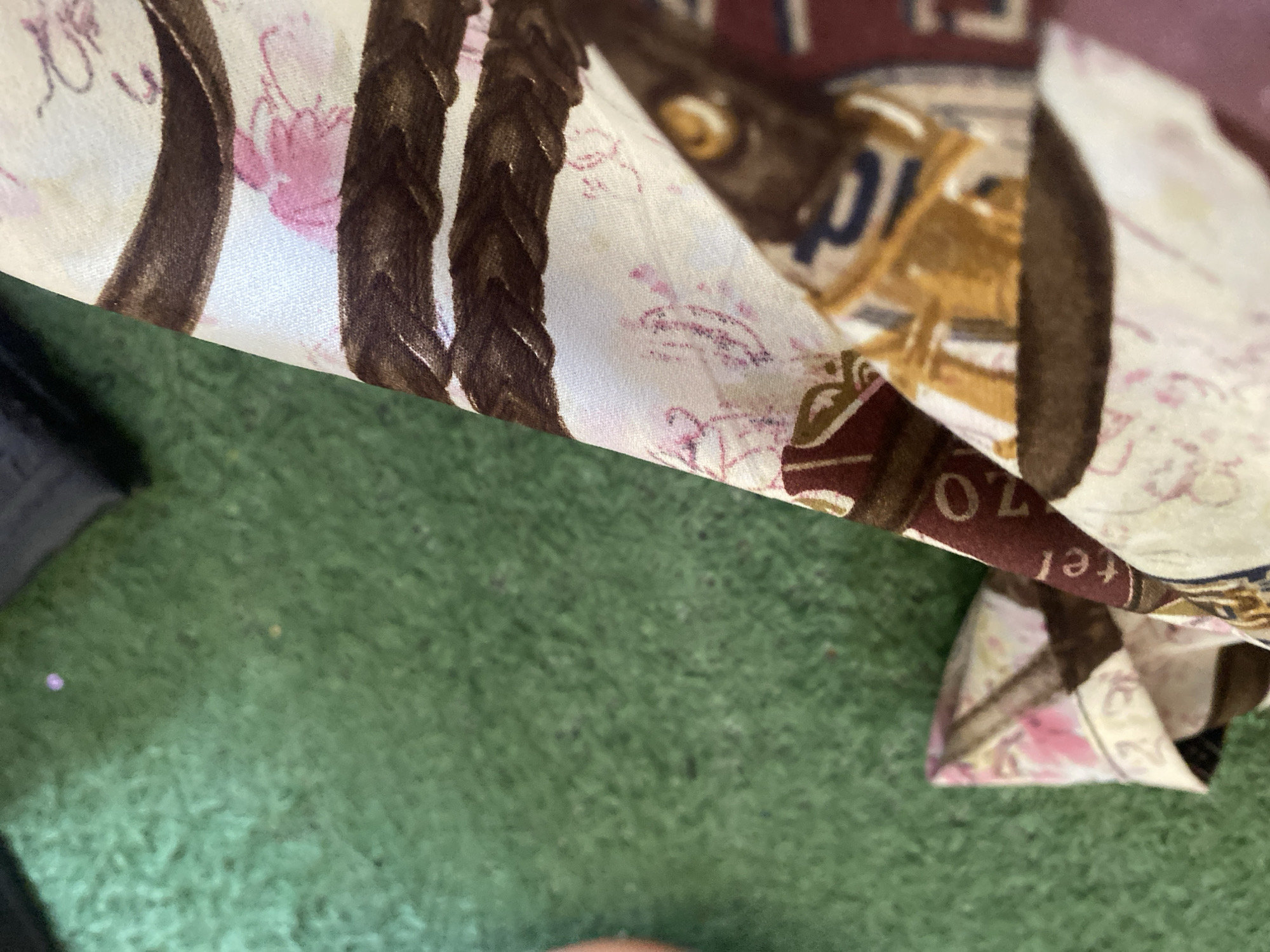 The image shows a close-up of a patterned fabric against a green, blurry background that appears to be a surface like artificial turf or carpet. The fabric has a design with brown stripes, pink floral elements, and some text that is partially visible. The text on the fabric includes the letters "tel" and the number "70". The overall scene is abstract due to the focus on the fabric. No specific location in Jauro Yinu, Nigeria can be identified from this image.