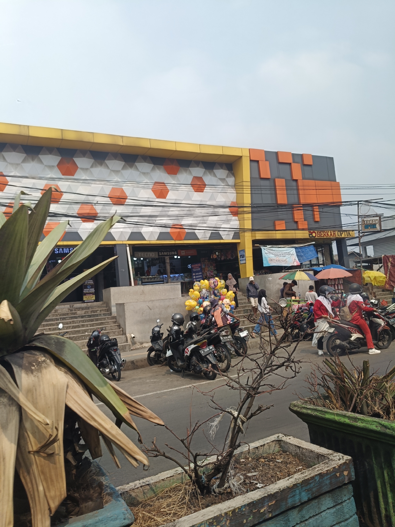 This image depicts a street view in Pandeglang, Indonesia, featuring a modern building with a distinctive yellow, orange, and gray hexagonal facade. The building appears to house several businesses, including "SAMSUNG" and "BERKAH OPTIK." In front of the building, there is a street with parked motorcycles and several individuals present. To the left, a large tropical plant with broad, dry leaves is prominent in the foreground, situated in a planter. Steps lead up to the entrance of the building, where a cluster of colorful balloons is visible. People are seen walking on the steps and in front of the shops. Further down the street, more motorcycles are parked, and a few people are on or dismounting from them. The environment suggests a busy commercial area on a clear day.