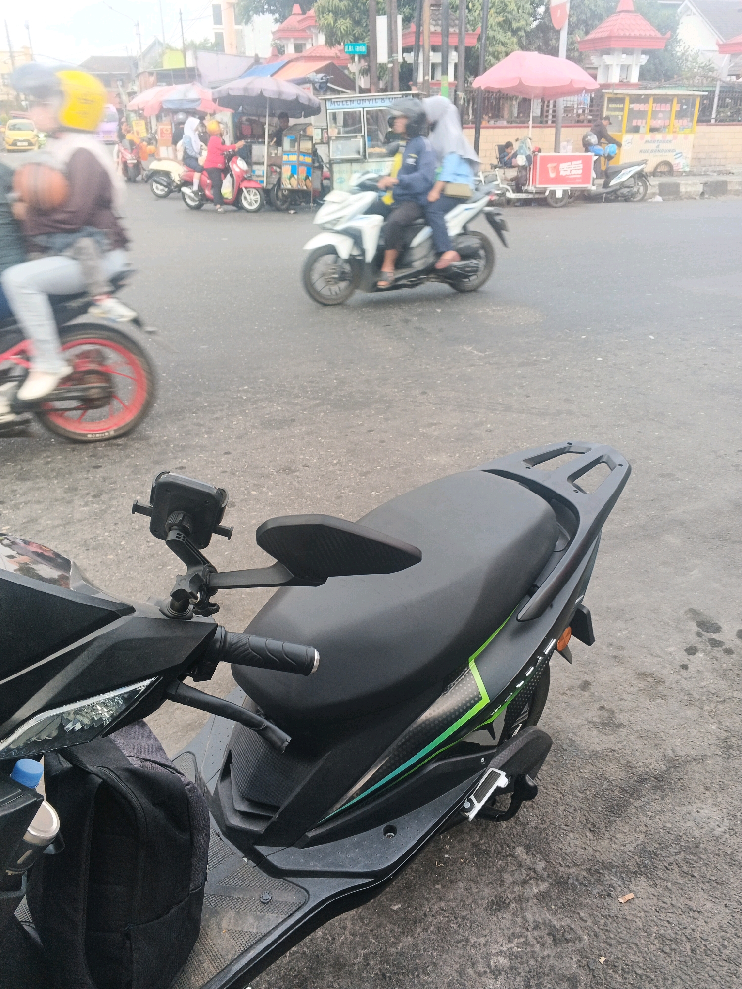 In Ungaran, Indonesia, a street scene unfolds with several motorbikes and a food cart. In the foreground, a black motorbike is parked, its front section featuring a phone mount and a black backpack. Further back, a white motorbike carries two individuals. To the left, another motorbike is in motion, with a person wearing a yellow helmet. In the background, a food cart with red and blue awnings serves customers, and other motorbikes are visible. The setting appears to be a busy urban area, possibly a street market or intersection.