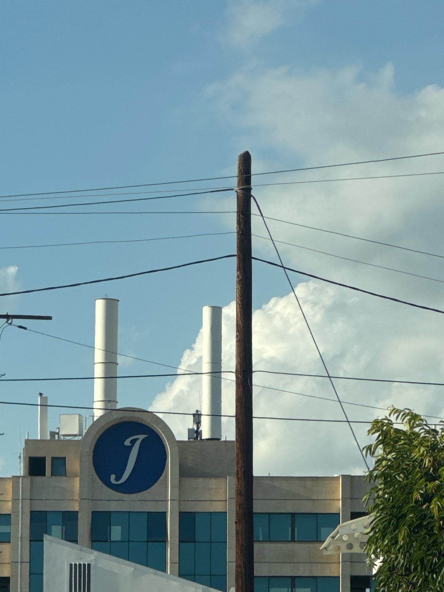 The image shows a building with a large blue circular logo featuring a white stylized letter "J." The building has a modern architectural style with multiple floors and large blue-tinted windows. Two tall, white cylindrical structures, possibly smokestacks or ventilation shafts, rise from the top of the building. A weathered wooden utility pole with several overhead wires stands in the foreground, obscuring parts of the building and sky. The sky is a light blue with scattered white clouds. A green leafy tree is visible on the right side of the image, partially obscuring the building. The scene appears to be an outdoor urban or industrial setting. Based on the provided information, this location is in the city of Mesa Geitonia, Cyprus.