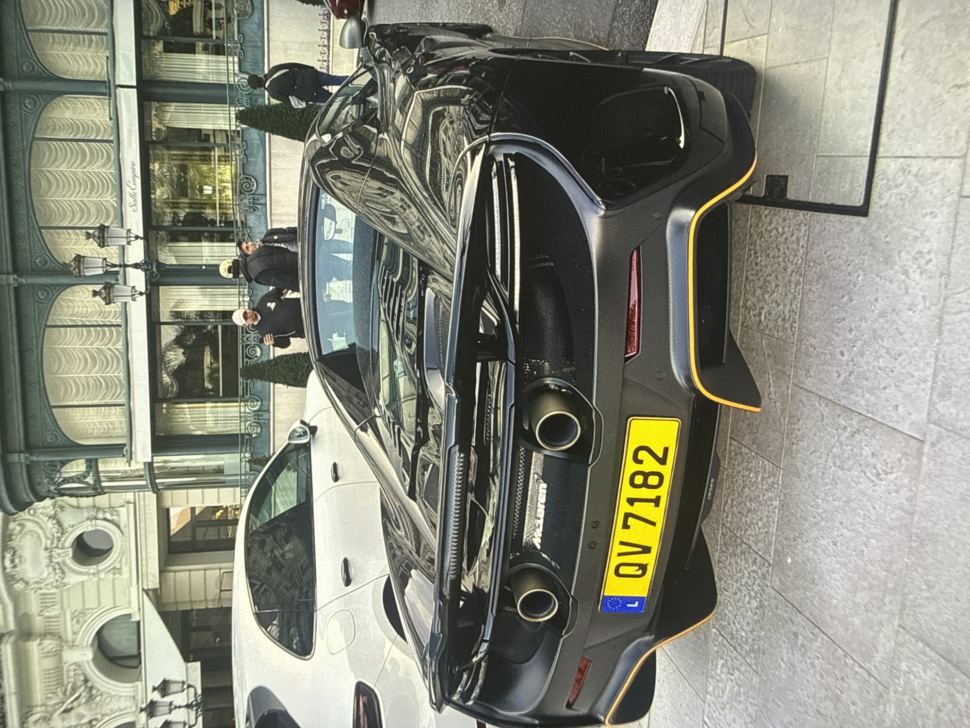 The image shows a black McLaren sports car parked on a paved surface. The car has a yellow Luxembourg license plate with the number "QV 7182". Next to the McLaren is a white car. Behind the cars, three people are standing in front of a building. The building has arched windows and a sign that says "Salle Empire". The image is not in Sofia, Bulgaria, and the location is most likely Monte Carlo.