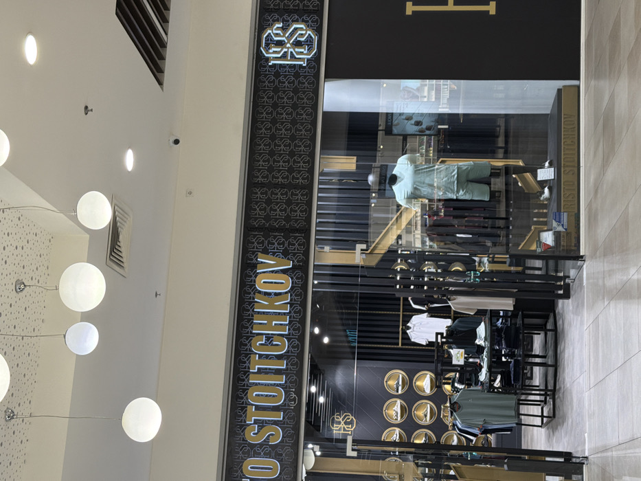 The image depicts the storefront of a retail clothing store named "RISTO STOITCHKOV" located within an indoor shopping mall in Stara Zagora, Bulgaria. The store's primary signage prominently displays "RISTO STOITCHKOV" in gold lettering with a light blue outline on a dark background featuring a repeating "HS" monogram pattern. A larger, stylized "HS" logo is also present on the right side of the main sign and above the entrance.
The storefront features a clear glass entrance and display windows framed by dark vertical structures with gold accents. Inside the right-hand display window, a headless mannequin wears a light teal short-sleeved t-shirt and matching shorts, with two pairs of white sneakers positioned on a dark display platform below. Visible through the glass on the left, various clothing items are displayed on hangers and folded on tables. A distinct display wall features multiple circular, gold-rimmed niches, each showcasing a single pair of athletic footwear. Other clothing items, including white and beige polo shirts, are presented on torso forms. The interior design incorporates dark vertical panels with gold-colored trim.
The mall's common area floor, composed of light-colored, polished rectangular tiles, extends to the store's threshold. The white ceiling above the storefront features a textured, light grey dotted pattern in one section, from which five spherical white pendant lights are suspended at varying heights. Additional recessed circular spotlights and rectangular air conditioning vents are visible on the ceiling. A small, black dome-shaped security camera is mounted on the ceiling, directed towards the storefront. No individuals are visible in the scene.