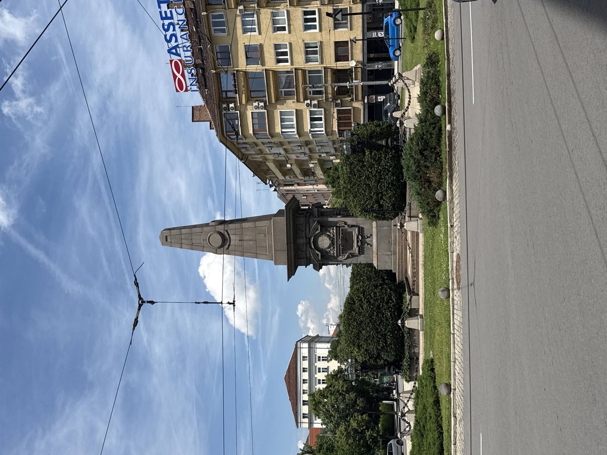 This image depicts a vibrant street scene in Sofia, Bulgaria, under a bright, sunny sky with scattered wispy clouds. The perspective is slightly angled, looking down a city street flanked by buildings and a central monument.
In the mid-ground, a tall, grey stone monument, resembling an obelisk, stands prominently. Its base is adorned with intricate carvings, including a circular wreath design, and is surrounded by stone posts connected by heavy chains. This monument is situated within a green, landscaped area featuring neatly trimmed bushes and trees.
To the right of the monument, a paved city road extends into the distance, marked by a single white line. Along the left edge of this road, a narrow grassy median dotted with spherical stone bollards separates the traffic from the monument's park-like setting. A blue car is visible parked on the far side of the street, while other vehicles are partially obscured further down.
The background is dominated by urban architecture. A multi-story building with a yellow or ochre facade stands to the right, characterized by numerous windows and air conditioning units. At the top of this building, a large sign clearly displays the text "ASSET INSURANCE" in red and blue letters, accompanied by a logo resembling an infinity symbol or a stylized '8'. Further down the street, another building with a lighter facade and a distinctive reddish-brown tiled roof is visible.
Overhead wires, likely part of a trolleybus or tram system, crisscross the blue sky. No people are distinctly visible, and the scene conveys the calm ambiance of a city center during daytime.