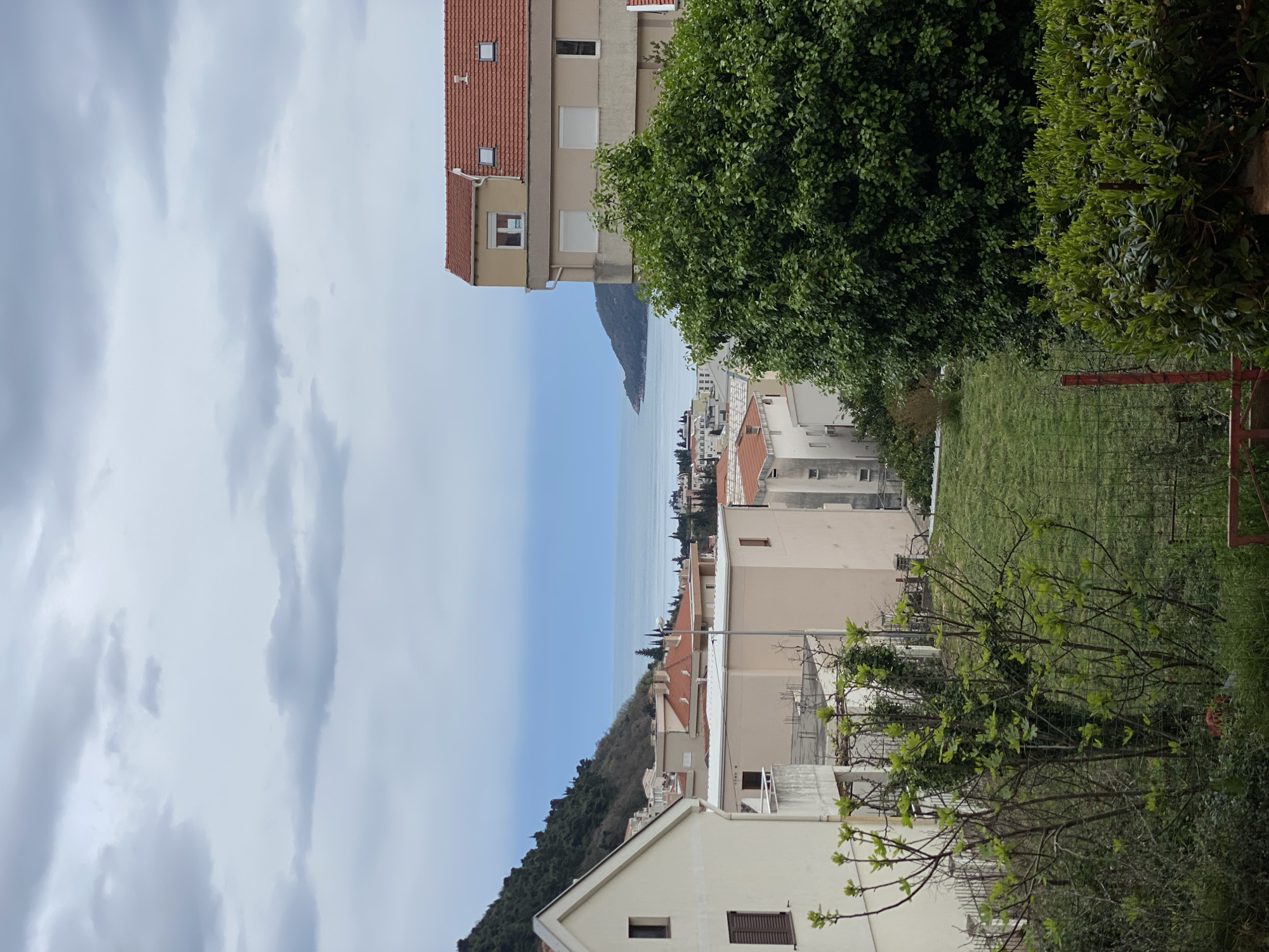The image shows a view of Budva, Montenegro. The scene is oriented vertically. Buildings are visible, ranging in height and architecture, with a mix of beige and white exteriors and red tiled roofs. The Adriatic Sea is seen in the distance, with a strip of land jutting out into the water. Overcast sky fills the left part of the frame. Trees and greenery are visible on the right side of the image. A small grassy area is framed by a chain-link fence and scattered vegetation.