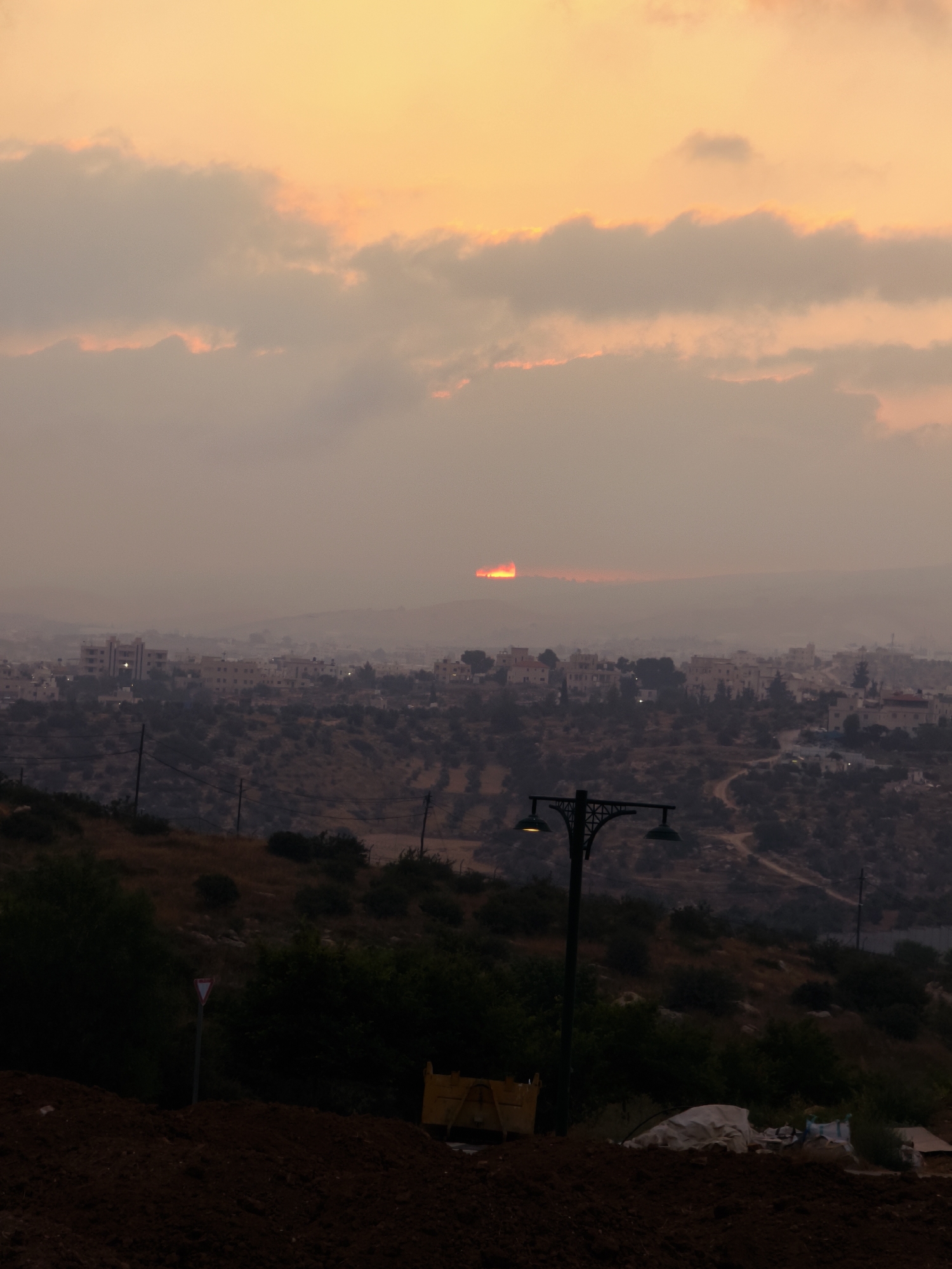 The image depicts a hazy sunset over the city of Lachish Regional Council, Israel. The sky is a gradient of orange and yellow, with clouds obscuring the setting sun, which is visible as a bright, distorted disk near the horizon. Below the sky, a densely populated town with light-colored buildings is spread across rolling hills. The foreground is dominated by a dark, dusty embankment, with a vintage-style streetlamp visible on the left. A yield sign is also present. The landscape is characterized by dry, scrubby vegetation and winding dirt roads. A yellow dumpster is partially visible on the right side of the image. The overall atmosphere is serene and slightly muted due to the haze.