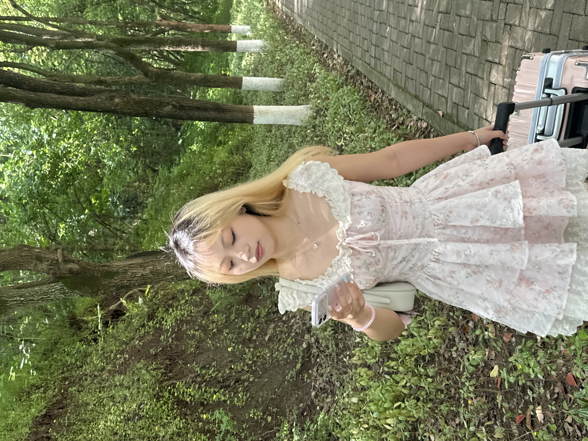A young woman with long blonde hair, featuring darker roots, is depicted outdoors, likely walking or paused on a paved path in Jiangjin, China. She is looking down intently at a white smartphone held horizontally in her hands. Her eyes appear closed or mostly closed, suggesting focus or a relaxed moment. She wears a light pink or white floral print dress with delicate ruffles around the neckline and shoulders. A light-colored handbag is slung over her shoulder, and she has a pink beaded bracelet on her right wrist, with light blue nail polish visible on some fingers. A delicate heart-shaped pendant hangs from her necklace.
To her right, a light pink suitcase with silver hardware and an extended handle stands on the paved surface, indicating travel. The paved path, made of gray rectangular blocks, runs alongside a grassy area abundant with green foliage and smaller plants. Several trees with prominent brown trunks and lush green leaves line the path. Notably, the bases of some tree trunks are painted white, a distinctive feature in the landscape. The scene is illuminated by natural daylight, suggesting it is daytime with bright, possibly diffused, sunlight. The overall setting appears to be a park or a well-maintained treelined area. No discernible text is visible in the image.