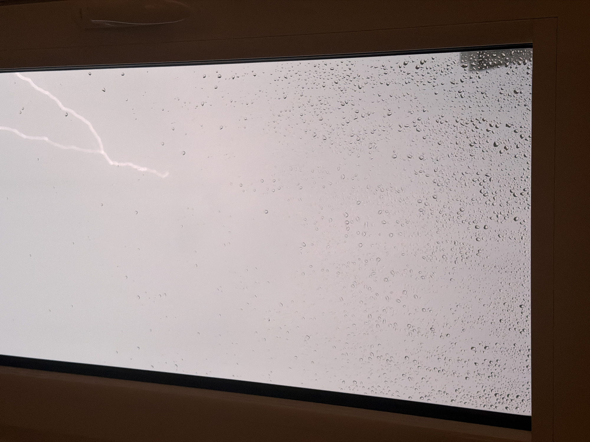 The image captures an indoor view looking out through a window during a thunderstorm. The windowpane is densely covered with numerous water droplets of varying sizes, indicating heavy rainfall. Through the wet glass, an overcast, light grey sky is visible, dominating the background.
A striking, bright white lightning bolt streaks across the upper left portion of the sky, forming a jagged, branching pattern. The lightning appears as a sharp, illuminated crack against the muted sky, highlighting the intensity of the weather event.
The window is framed by a dark brown or beige border, visible at the top, bottom, and right sides of the image. This frame suggests the view is from inside a building, looking out. The arrangement implies the window might be a skylight or a high-set window, given the angle and the visible upper frame.
The scene conveys the drama of a natural event – a rainstorm with lightning – viewed from a protected interior space. No people or specific objects are visible indoors, keeping the focus on the stormy exterior. The time of day appears to be daytime, though heavily clouded by the storm. There is no visible text in the image. This meteorological event is occurring in Budva, Montenegro.