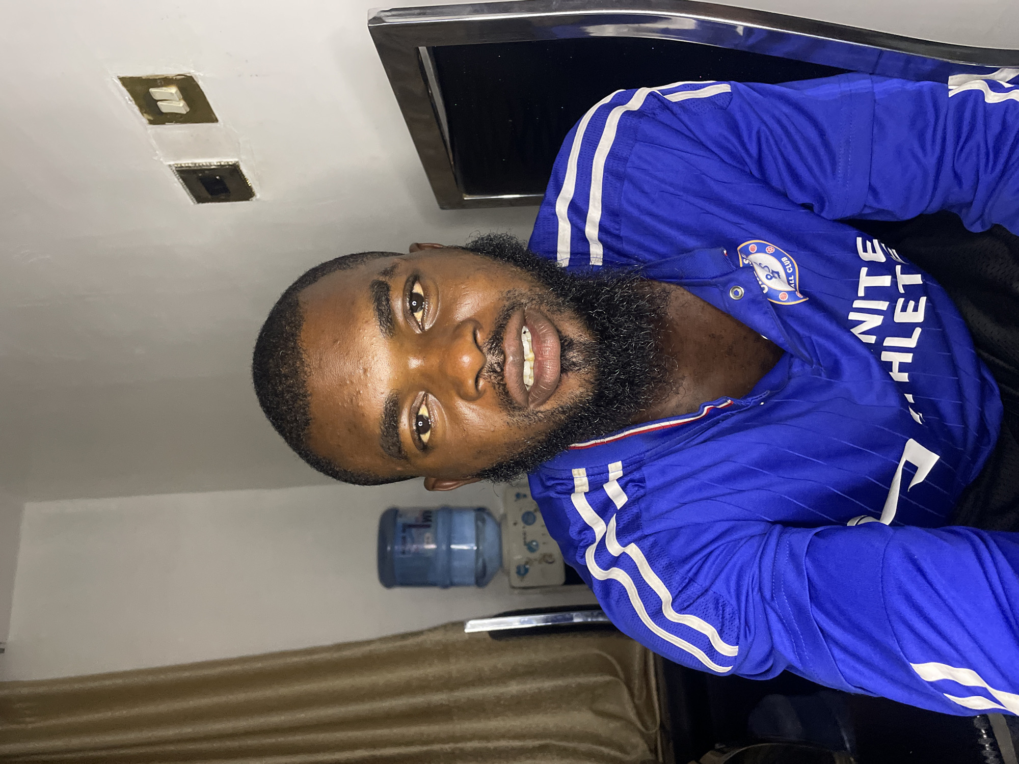 The image shows a medium shot of a Black man with a beard, wearing a blue jersey with white stripes on the sleeves. He is looking directly at the camera with an open mouth. The man is seated indoors, with a water cooler visible behind him to the left. To the right, a metallic frame, possibly part of a piece of furniture or a screen, is present. The background wall appears to be painted white, with two light switches or electrical outlets. The lighting suggests an indoor setting, possibly a home or office. The man's attire indicates he might be a sports enthusiast or affiliated with a team. The overall impression is a casual portrait taken within a domestic or functional indoor environment. There is no indication of the specific location in the city of Jos, Nigeria, beyond it being an indoor scene.