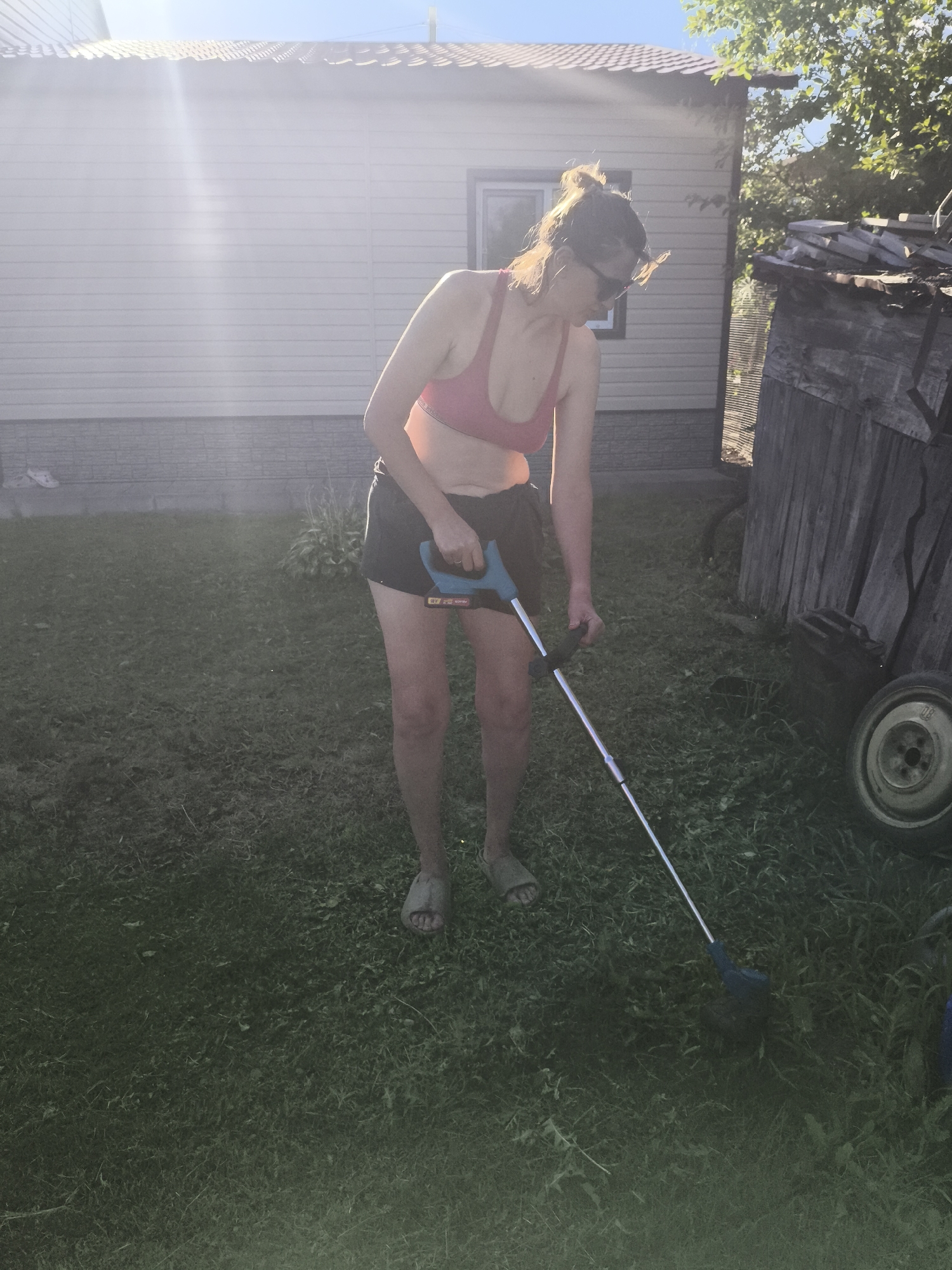 A woman is trimming grass with a weed trimmer in the yard of a rural home. The woman is wearing a pink sports bra and black shorts. She has her hair pulled up in a bun and is wearing sunglasses. She is holding the weed trimmer with both hands and is focused on the grass. The yard is covered in green grass and there are some weeds. In the background, there is a white house and a wooden shed. The sky is blue and clear. The location is the city of Babruysk, Belarus.