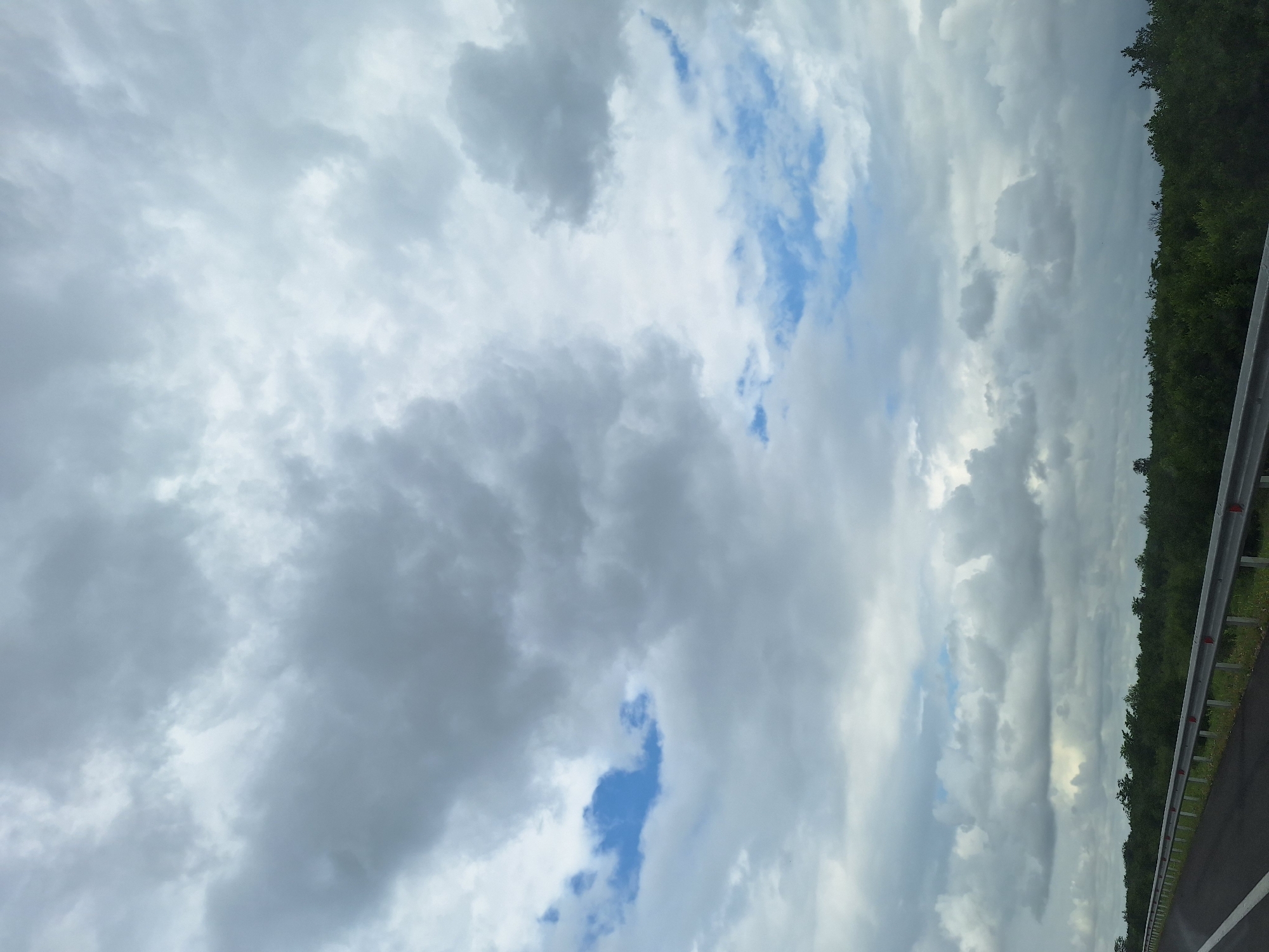 The image depicts a roadside view with a cloudy sky overhead. The foreground features an asphalt road with a white line marking the lane. A metal guardrail runs along the edge of the road. Beyond the road, a dense line of green trees forms the horizon. The sky is filled with a mix of grey and white clouds, with patches of blue sky visible in between. The scene appears to be a natural landscape, possibly in a rural or suburban area, given the presence of trees and the absence of urban structures. The lighting suggests it is daytime, but the cloud cover creates a somewhat muted atmosphere.
