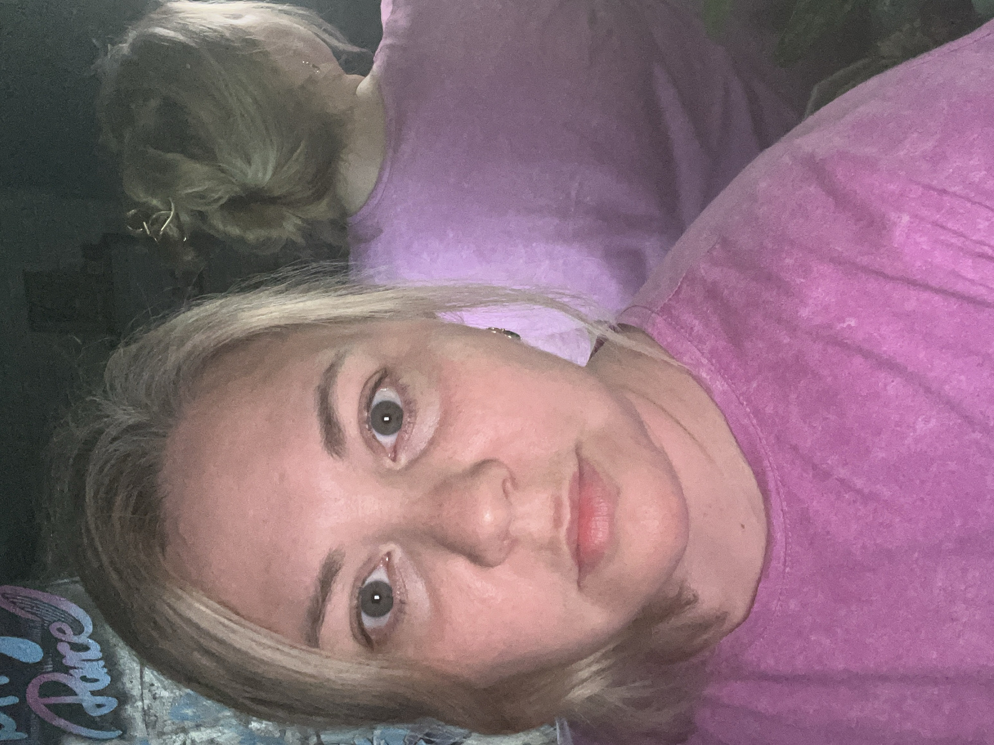 A selfie of a woman with blonde hair and fair skin wearing a pink t-shirt. Another woman with blonde hair is visible out of focus in the background, wearing a similar pink shirt. The background also contains a dark, undefined area and a patterned surface in shades of blue and purple. No location information is provided.