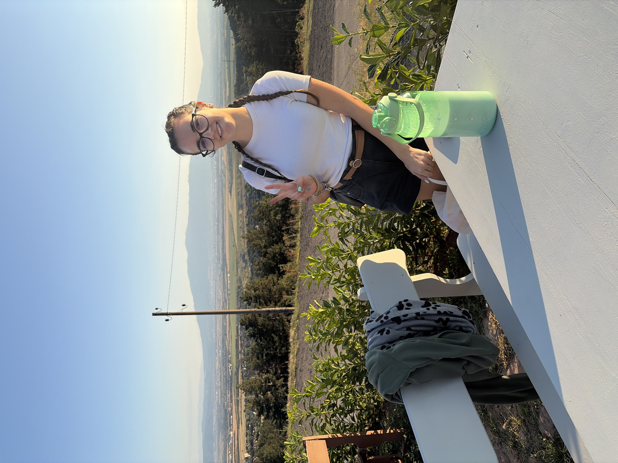 A young woman with dark hair in braids and glasses poses with a peace sign in front of a scenic overlook. She is wearing a white t-shirt, black shorts, and a brown belt. A green water bottle is on a white picnic table in the foreground, and a bench behind her has a green jacket and a patterned scarf draped over it. The background shows a wide valley with a city in the distance, rolling hills, and mountains under a clear blue sky. The setting appears to be a park or viewpoint on the outskirts of Sofia, Bulgaria, likely Vitosha Mountain, offering a panoramic view of the city and surrounding landscape.