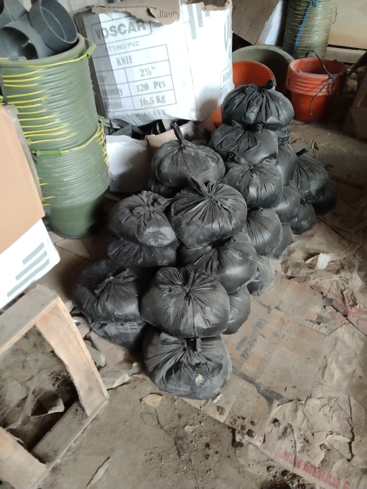 In Parung, Indonesia, a pile of black trash bags is stacked on the floor of what appears to be a storage area or warehouse. The bags are tightly bound at the top, obscuring their contents. Behind the trash bags, several large green cylindrical containers are visible, stacked on top of each other. To the right of the trash bags, orange buckets are stacked, and further back, a large white box with text that includes "VOSCAR" and "KNIE" is present. The environment is dusty and cluttered with various objects, including cardboard and what looks like part of a wooden crate or pallet in the foreground. The lighting is dim, suggesting an indoor, possibly unlit, space.