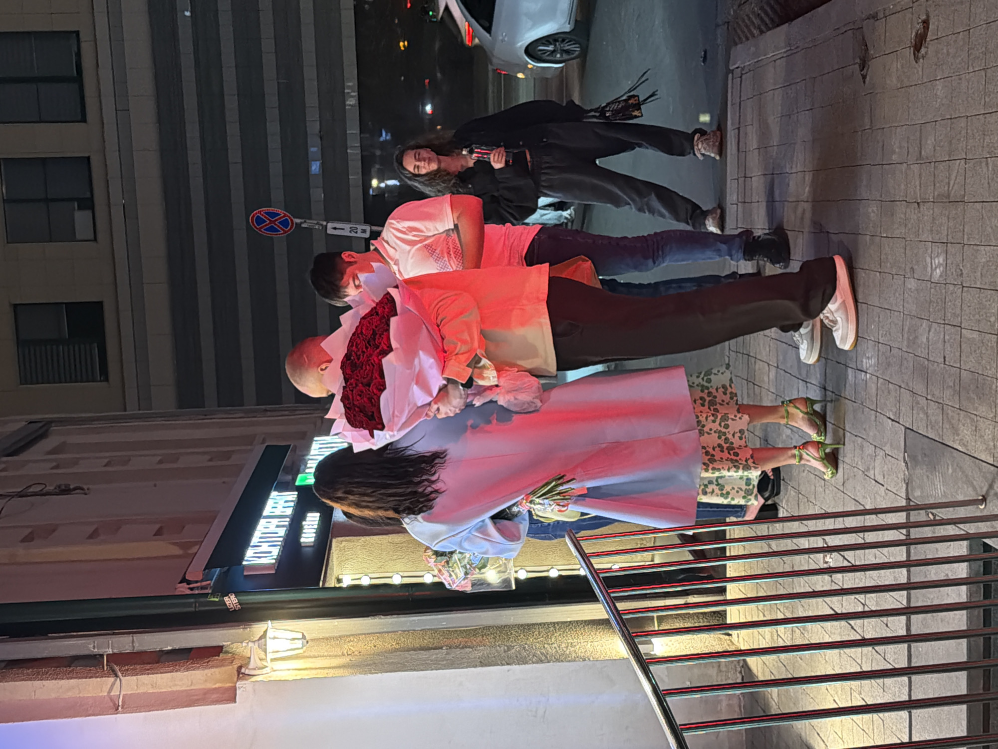 A group of people stands on a city street at night. A bald man holds a bouquet of red roses, and a woman in a light blue coat holds another bouquet. A woman in a black jacket holds a bottle. They stand on the sidewalk next to a building with a sign that reads "Khaiber Stereo." In the background, there is a street sign with an "X" on it. The location is identified as Moscow, Russia.