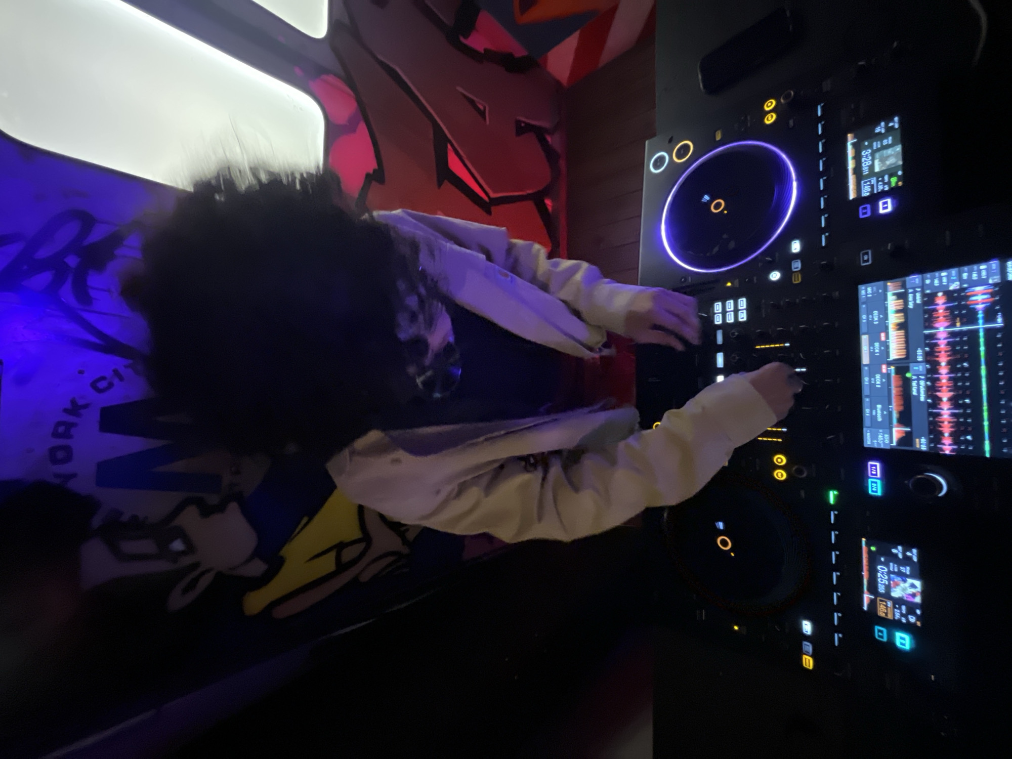 The image shows a person with dark hair and a light-colored jacket operating a Pioneer DJ controller, presumably in a nightclub or similar venue. The controller features a blue-lit jog wheel and multiple screens displaying waveforms and track information. In the background, there is graffiti on the wall. A pillow with "NEW YORK CITY" lettering and a cartoon graphic is also visible. The room is dimly lit. There's no evidence to determine this image was taken in Barcelona, Spain.