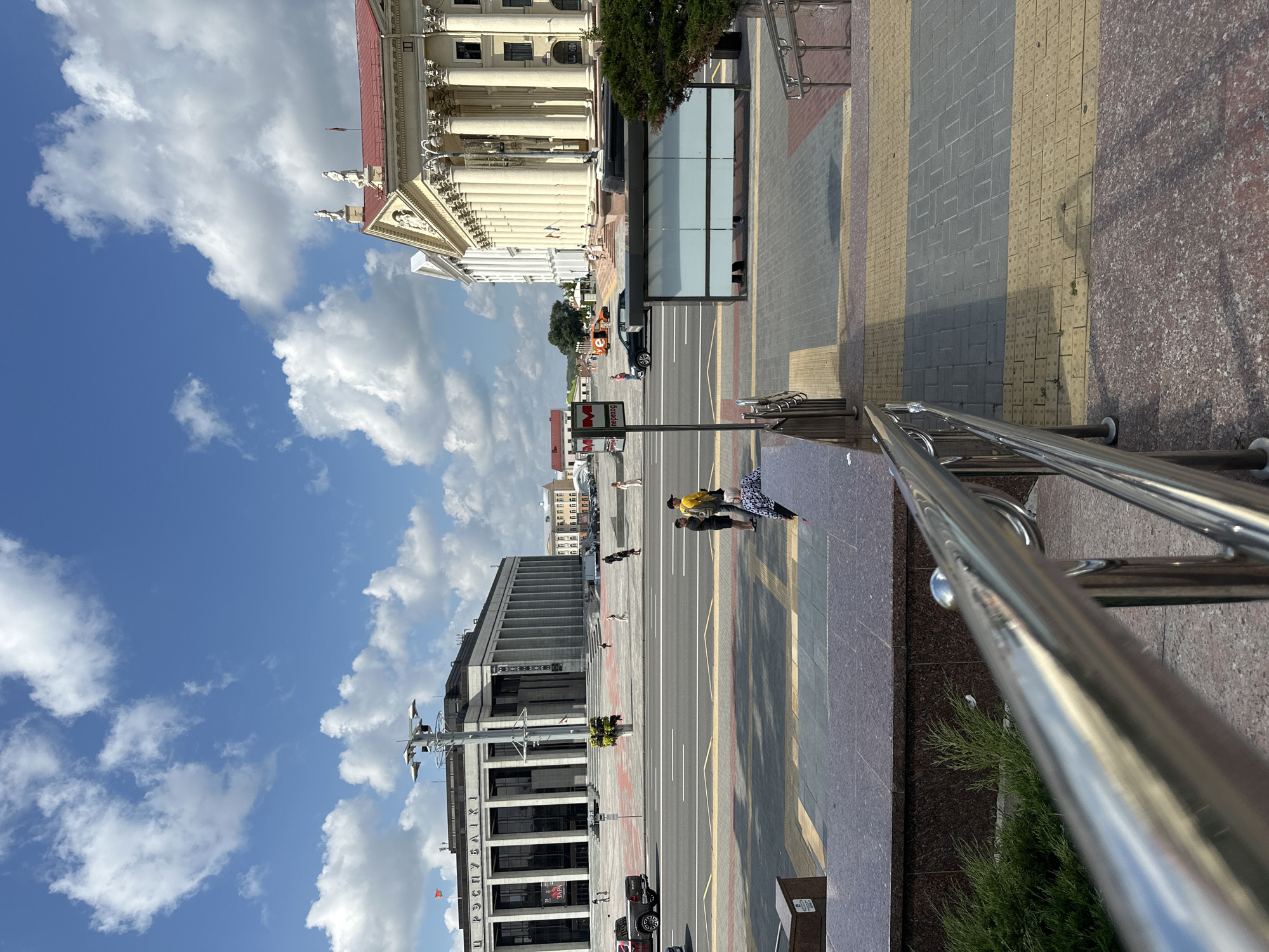 The image depicts a wide street scene in Minsk, Belarus, on a partly cloudy day. In the foreground, a polished metal handrail lines a set of stairs made of speckled granite and patterned paving stones. The stairs descend from the viewer's perspective, leading down to a pedestrian walkway.
Across the street, two prominent buildings dominate the view. On the left is a large, modern building with a colonnaded facade, likely a governmental or cultural institution. Cyrillic text is visible on its exterior. On the right stands a classical-style building with a red roof, ornate columns, and statues adorning its upper level.
Several people are visible on the sidewalk and street. Two individuals are standing on the sidewalk near a bus stop shelter. A few other pedestrians are scattered across the plaza in front of the large building on the left. Cars are present on the street, including a black SUV and a silver pickup truck. The sky is blue with scattered white cumulus clouds. The overall impression is that of a well-maintained urban public space on a bright, clear day.