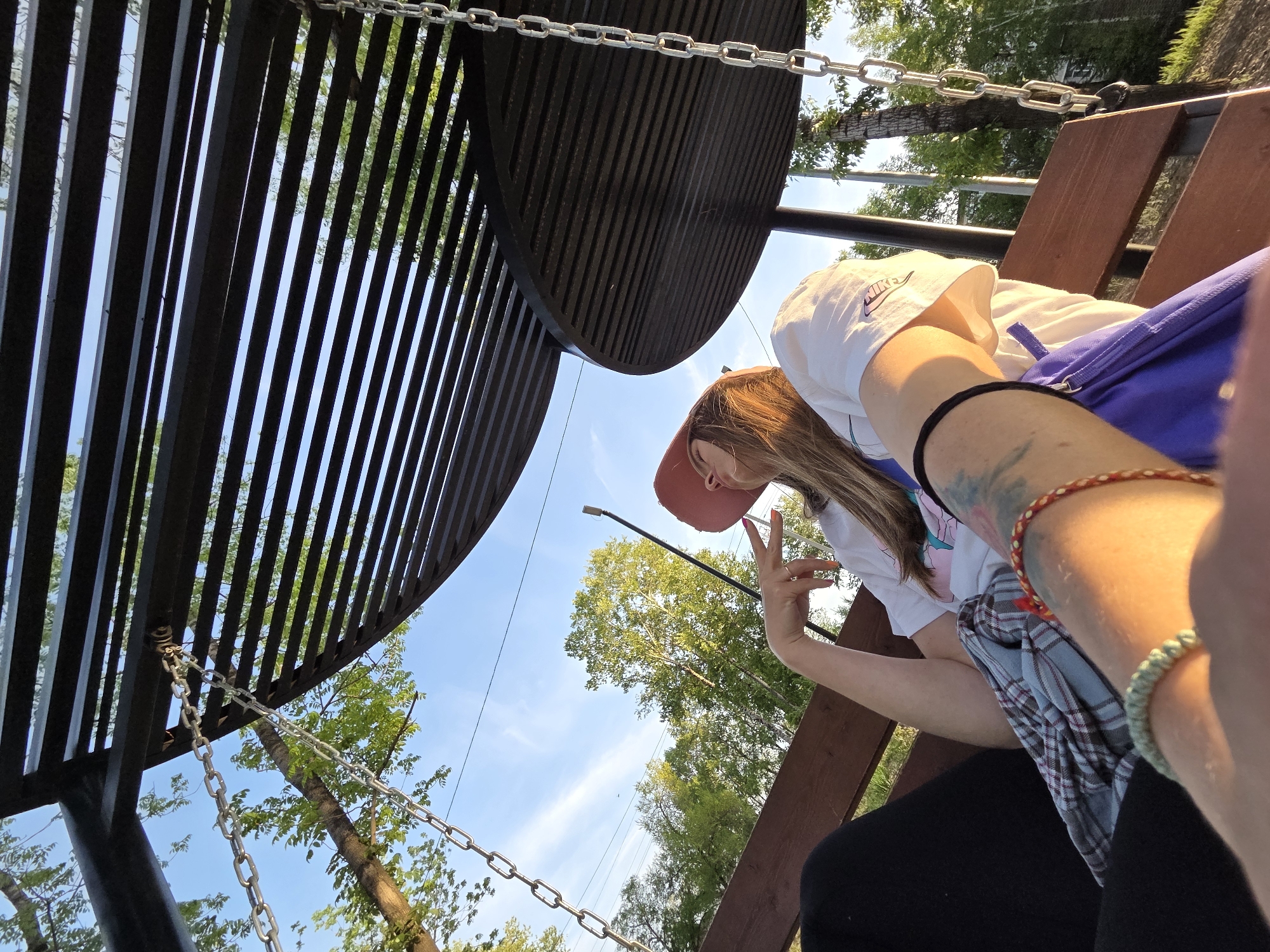 A low-angle, wide shot of a person sitting on a swing set. A woman with light brown hair wears a pink baseball cap, a white t-shirt, and black pants. She holds her hand up in a "peace" sign. The swing hangs from a metal structure. Trees and a blue sky are visible in the background. The location is outdoors, but no specific details indicate Birobidzhan, Russia.