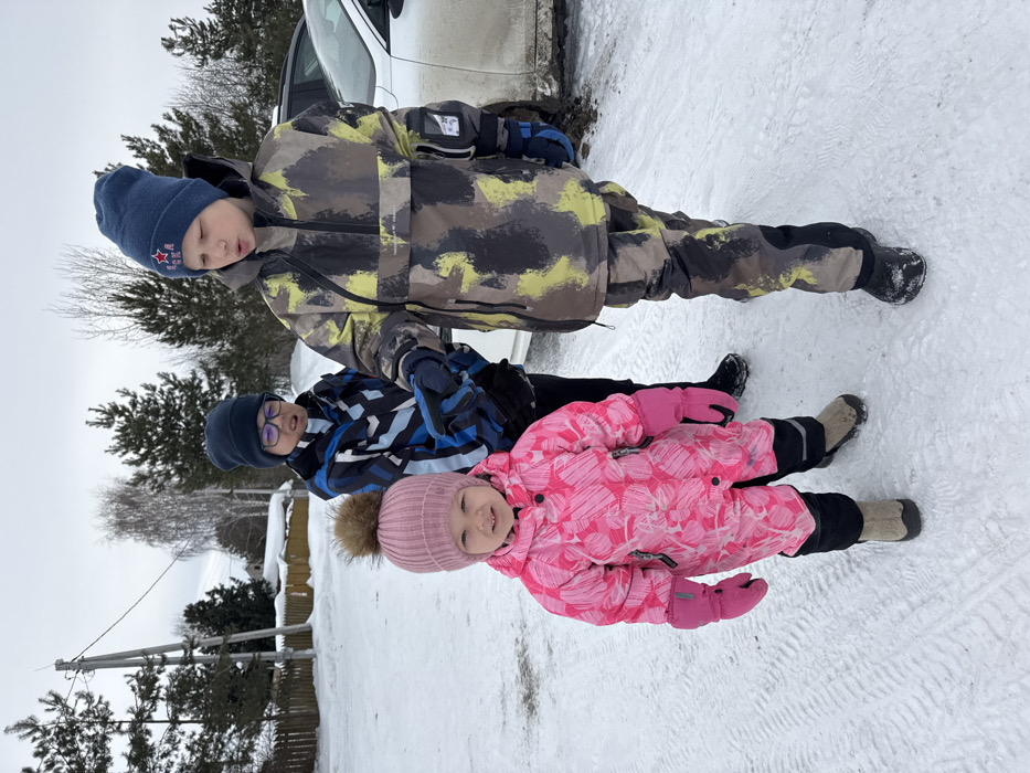 An outdoor scene captured in Zaosinovo, Russia, depicts three individuals, appearing to be children, standing on a snow-covered surface under an overcast sky.
From left to right:
1. A young child, approximately 3-5 years old, faces the camera with a slight smile. This child wears a pink ribbed knit hat with a brown fur pompom, a pink and white patterned full snowsuit, and pink gloves. Beige winter boots are visible.
2. Behind and slightly to the right of the first child, a second child, appearing older, stands looking forward with a slightly open mouth. This child wears a dark blue knit hat, dark-framed glasses, a blue, white, and black patterned winter jacket, and dark pants. The child's right hand is positioned near the third child's left arm.
3. To the right, a third child, appearing the oldest of the three, looks down and slightly to their left. This child wears a blue knit hat with a red star and "CK" embroidered on it, a camouflage-patterned (green, black, grey, yellow) winter jacket and matching pants, and dark blue gloves. A black object, possibly a smartphone or small device, is held in the child's left hand.
The ground is covered in packed snow, exhibiting faint tire tracks in the foreground. In the background, a white car is partially visible on the right. Further back, there are wooden fences, utility poles with wires, snow-covered structures consistent with houses, and a mix of bare deciduous trees and green coniferous trees. The sky is uniformly grey and overcast. All individuals are dressed in heavy winter clothing suitable for cold conditions.