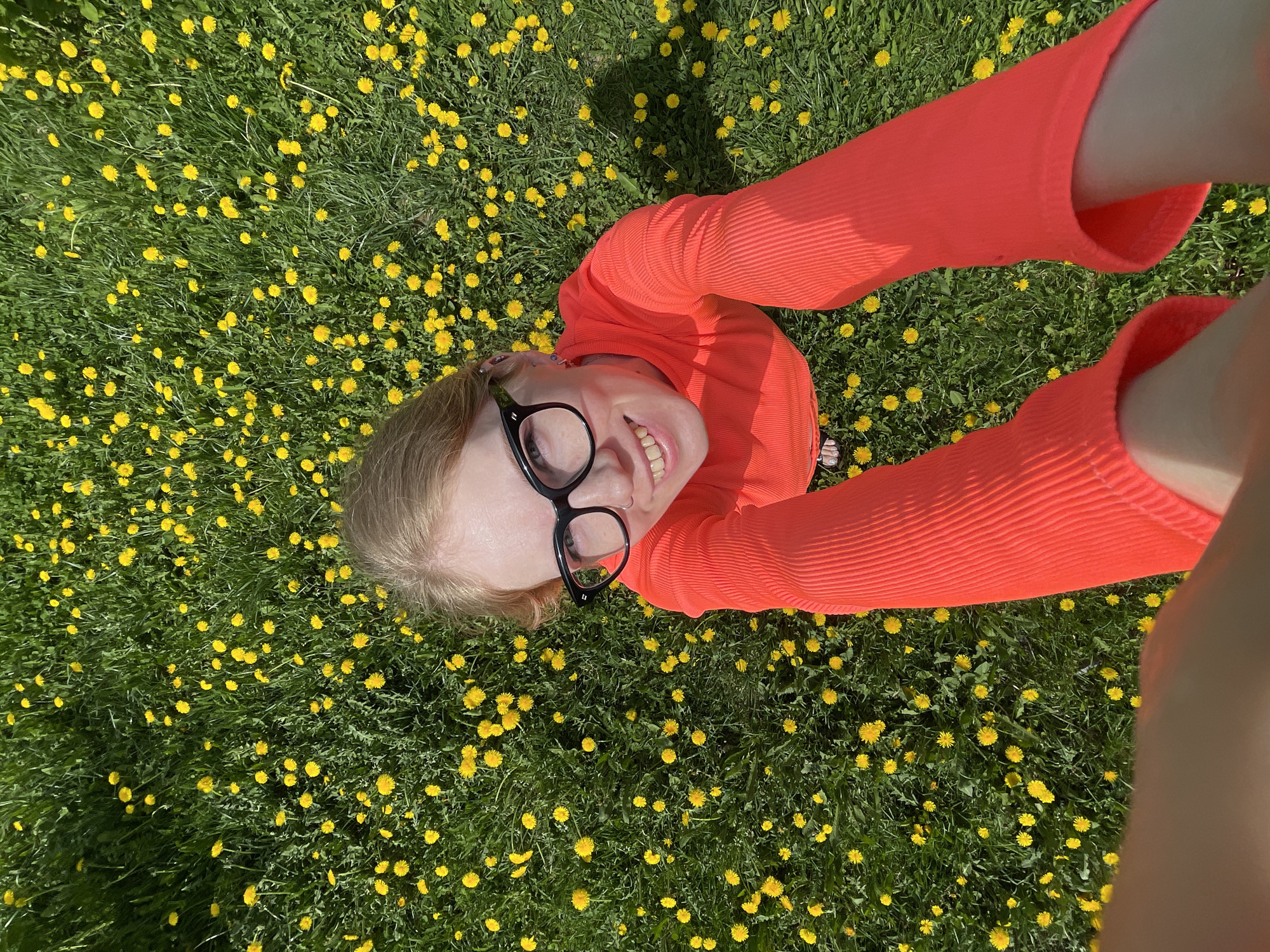 A young woman lies on her back in a field of dandelions, smiling up at the camera. She wears large black glasses and a bright orange, long-sleeved shirt. The location appears to be outdoors in a grassy area covered with yellow dandelions. The photograph seems to be taken at a close distance, suggesting she might be taking a selfie. No information indicates the location is Izhevsk, Russia.