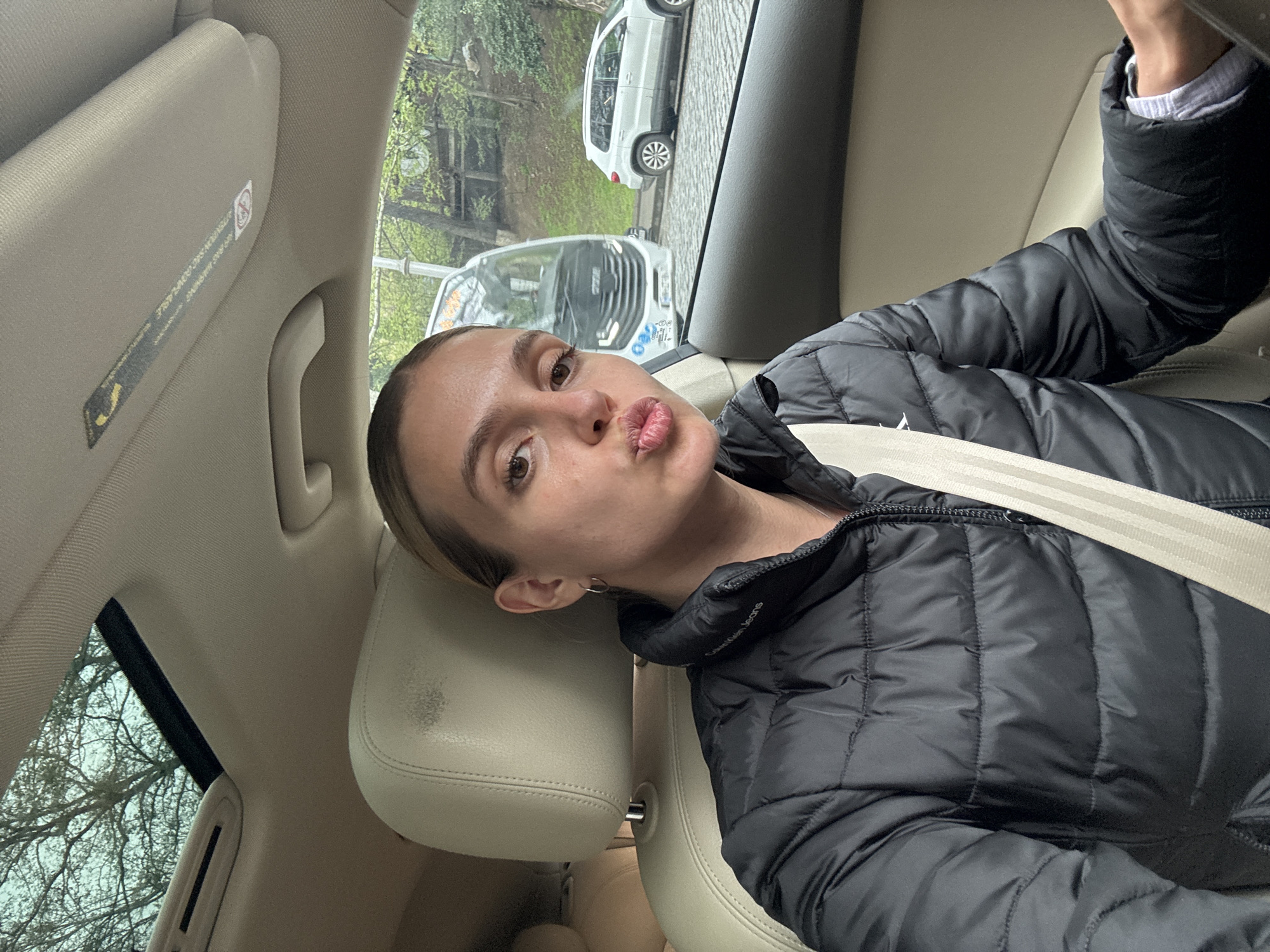 A young woman is captured in a selfie or photo inside a car, looking towards the camera with a playful "duck lips" pout. She has light skin, dark eyes, and her hair is pulled back. She is wearing a black puffer jacket with "Calvin Klein Jeans" visible on the sleeve, over a light-colored shirt. A beige seatbelt crosses her chest, and she is seated in a beige leather car interior, with a headrest and sun visor visible.
Through the car window, a city street in Tbilisi, Georgia, is visible. The street appears to be paved with cobblestones. Several vehicles are parked or moving outside, including a white SUV and a white van or minibus. The white van has text on its side and front, consistent with local transport in Tbilisi, Georgia. The trees outside have bare branches, suggesting an autumn, winter, or early spring setting. The sky appears overcast or partly cloudy, with diffused daylight indicating it is daytime.
Visible text on the car's sun visor includes "WARNING: TIRE PRESSURE MONITORING..." and a yellow safety symbol. The woman's jacket clearly shows the "Calvin Klein Jeans" logo on the left sleeve.