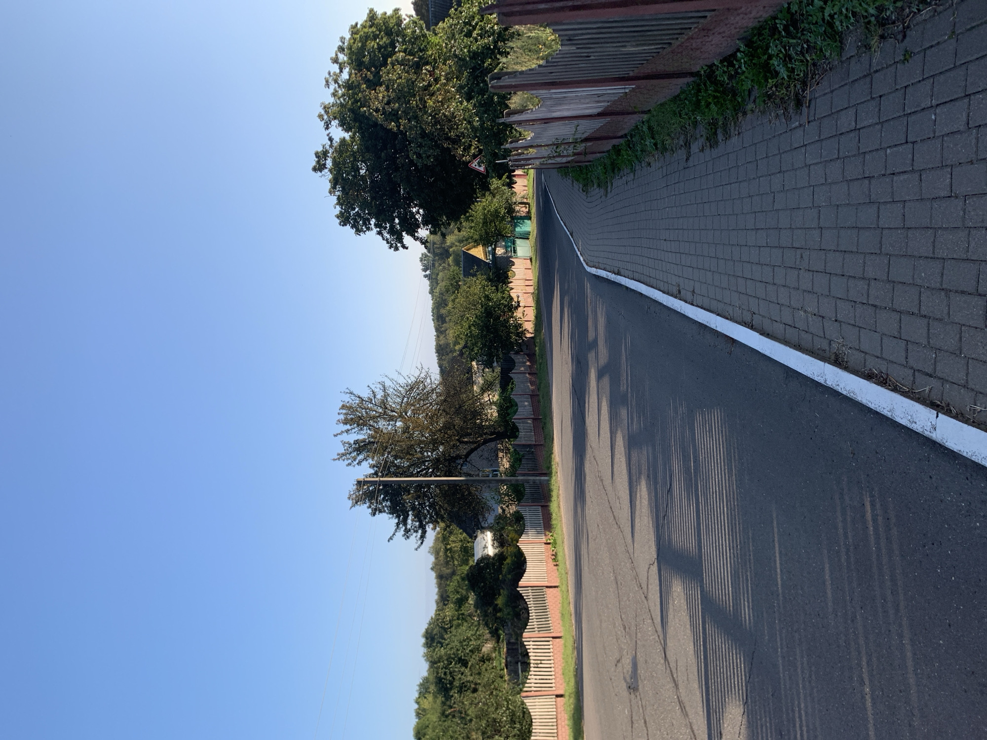 The image depicts a quiet, rural road in the city of Kapyĺ, Belarus, under a clear blue sky. The road, paved with asphalt, curves to the right, bordered by a cobblestone sidewalk on the right and a wooden picket fence on both sides. The fence is painted a reddish-brown, with lighter wooden posts. Shadows of the fence are cast across the road, suggesting it is late afternoon or early morning.
On the left side of the road, beyond the fence, are several houses with traditional gabled roofs, partially obscured by dense green trees and foliage. A lone utility pole stands near the center of the frame, with power lines stretching across the sky. On the right side, the fence continues along the curving road, and beyond it, more trees and glimpses of houses can be seen. The overall atmosphere is tranquil and serene, characteristic of a village or small town setting. There are no people or animals visible in the image.