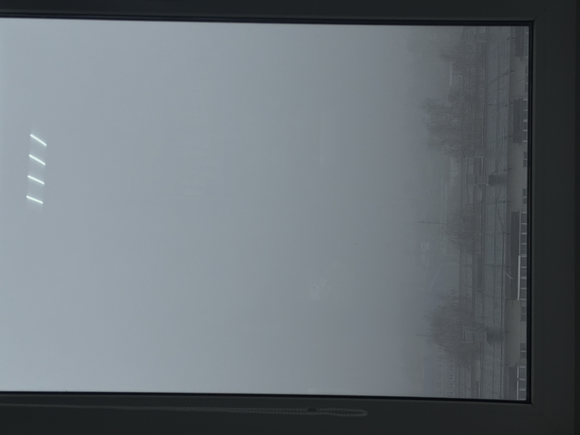 This image presents a static view through a dark-framed window, looking out onto a heavily fog-obscured landscape in Chaikovsky, Russia. The predominant color tone is a muted grey, reflecting the dense atmospheric conditions.
On the left side of the window, four bright, parallel, slightly angled white lines are clearly visible, appearing as reflections of interior fluorescent or LED lights from the room where the photograph was taken. The window pane itself shows some minor smudges or imperfections in the center.
Beyond the window, the exterior scene is enveloped in a thick fog or mist, severely limiting visibility. Faint, blurred outlines of what appear to be multi-story buildings, possibly residential or industrial structures, and bare trees can be discerned on the right side of the frame, emerging like shadows from the grey haze. The architectural style, though indistinct, aligns with urban environments found in the region.
The weather is characterized by heavy fog and extremely overcast skies, creating a dim and diffused daytime atmosphere. No specific activities or people are visible due to the poor visibility. The overall mood conveyed is one of quietude and obscurity. No text is visible in the image.