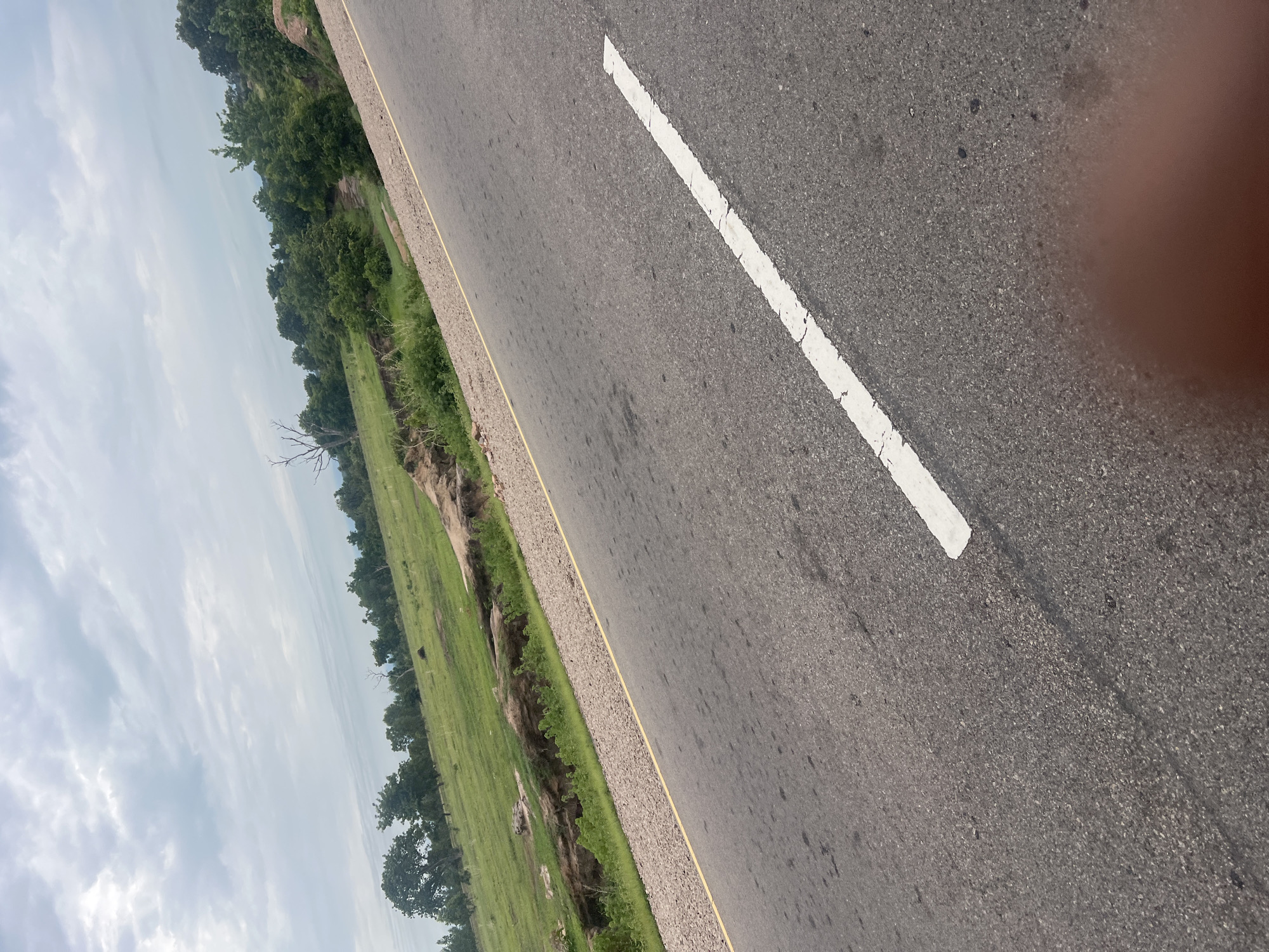 The image depicts a paved road angling across the frame from the lower right to the upper left. A white line marks the center or lane division of the road. To the left of the road, a green, grassy landscape slopes upwards, featuring scattered trees, some with bare branches, and rocky outcrops. A ditch or erosion channel is visible in the grassy area. The sky above is overcast with pale clouds. A blurry reddish-brown area, possibly a thumb or finger, is visible in the lower right corner of the image. The scene appears to be a rural or semi-rural environment, consistent with the outskirts of a location like Minda, Nigeria, given the natural vegetation and road type.