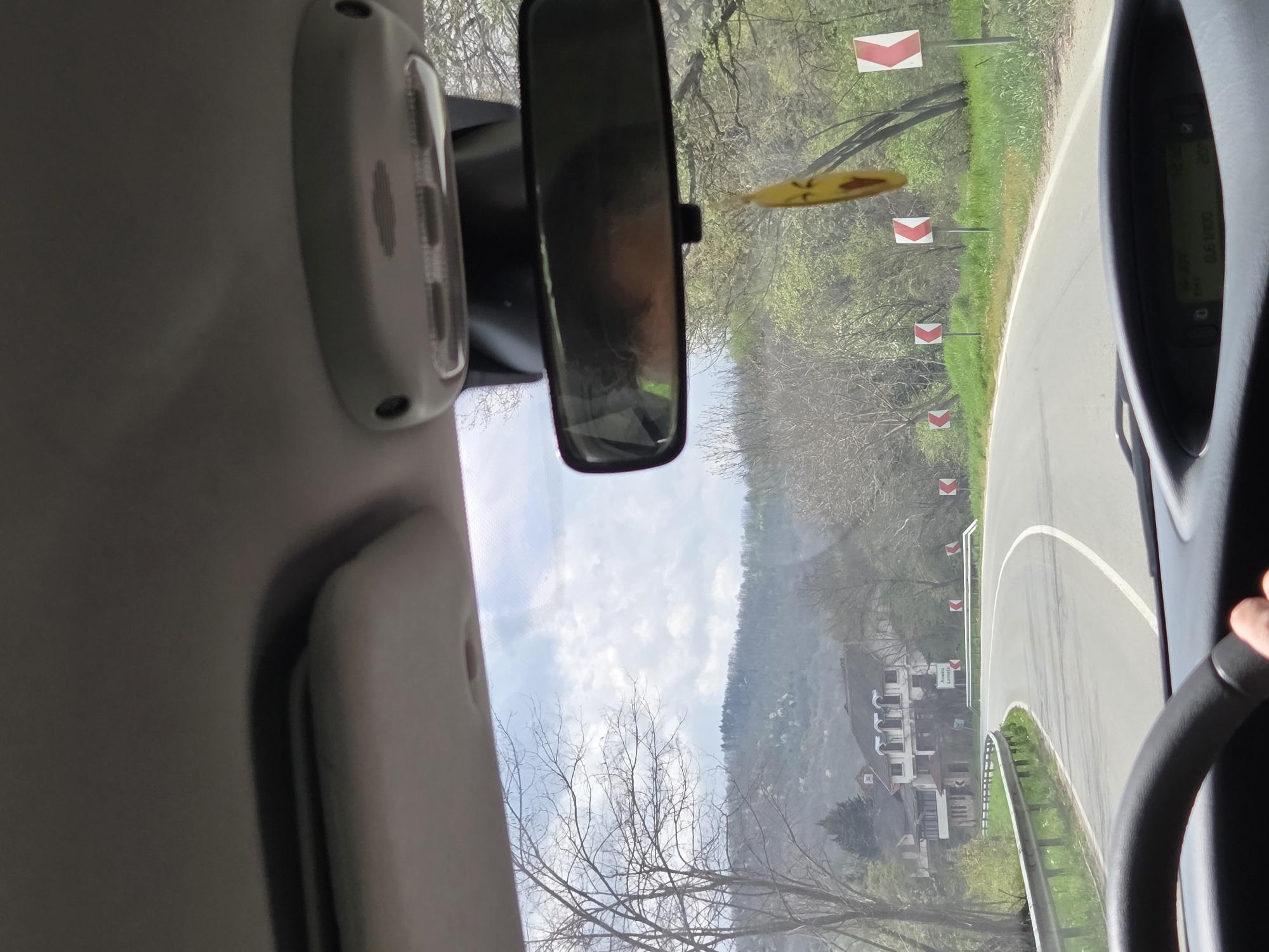 The view from inside a vehicle, driving on a winding road marked with red and white chevron signs. Outside, there are trees and a building with a sign indicating "Adamec Lomnica" visible, likely a hotel or business in the Leshnitsa, Bulgaria area. A driver's hand is visible on the steering wheel.