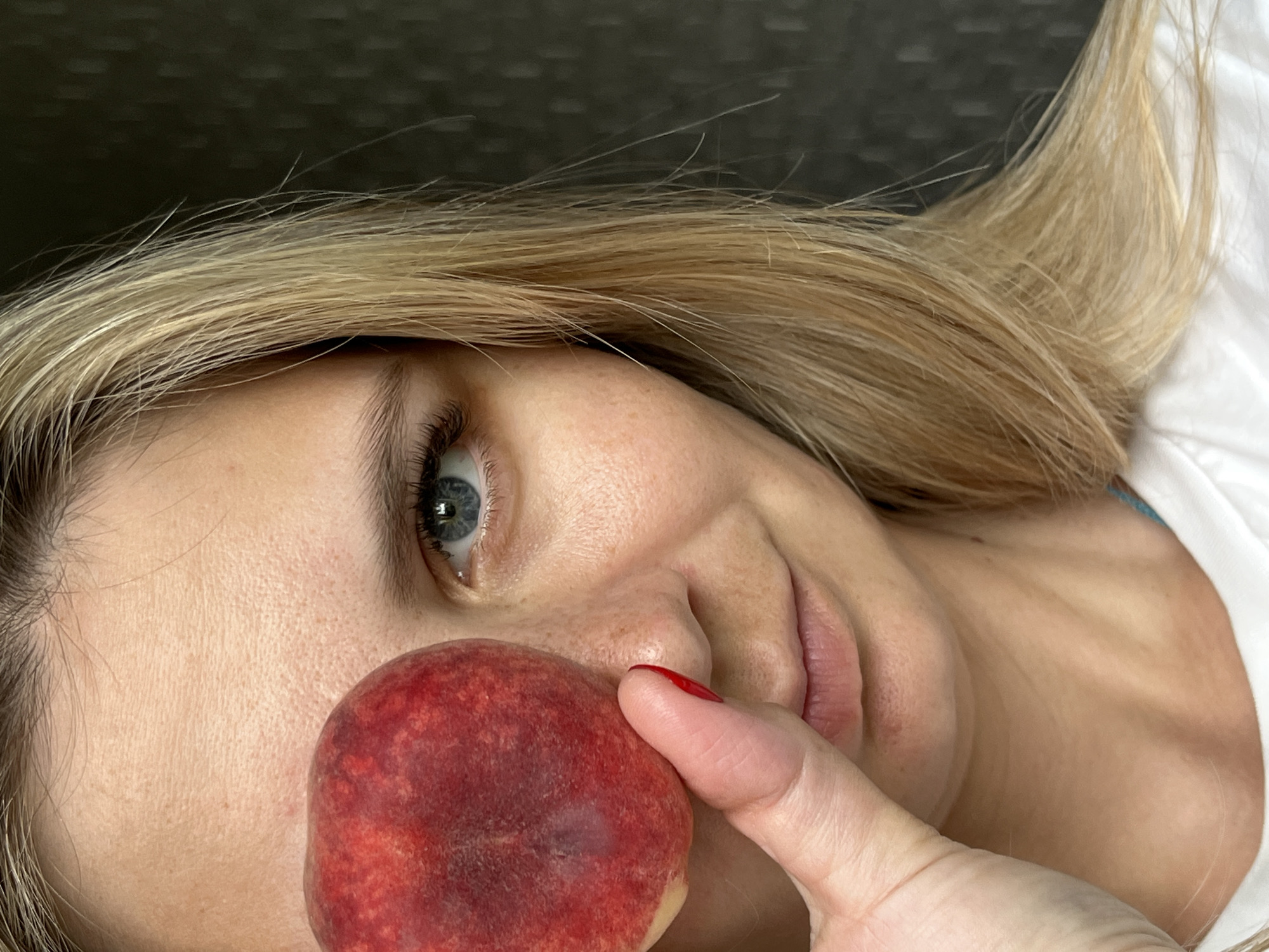 A close-up, shoulder-up shot shows the right side of a blonde woman's face. She is wearing a white t-shirt. Her right eye is visible and has blue irises. Her hair is blonde and falls around her face and shoulder. She is holding a red fruit, possibly a peach or nectarine, up to her face, covering her left eye. Her right hand is visible, with a red-painted fingernail. The background is a dark, textured surface. The image was taken in the city of Chaikovsky, Russia.