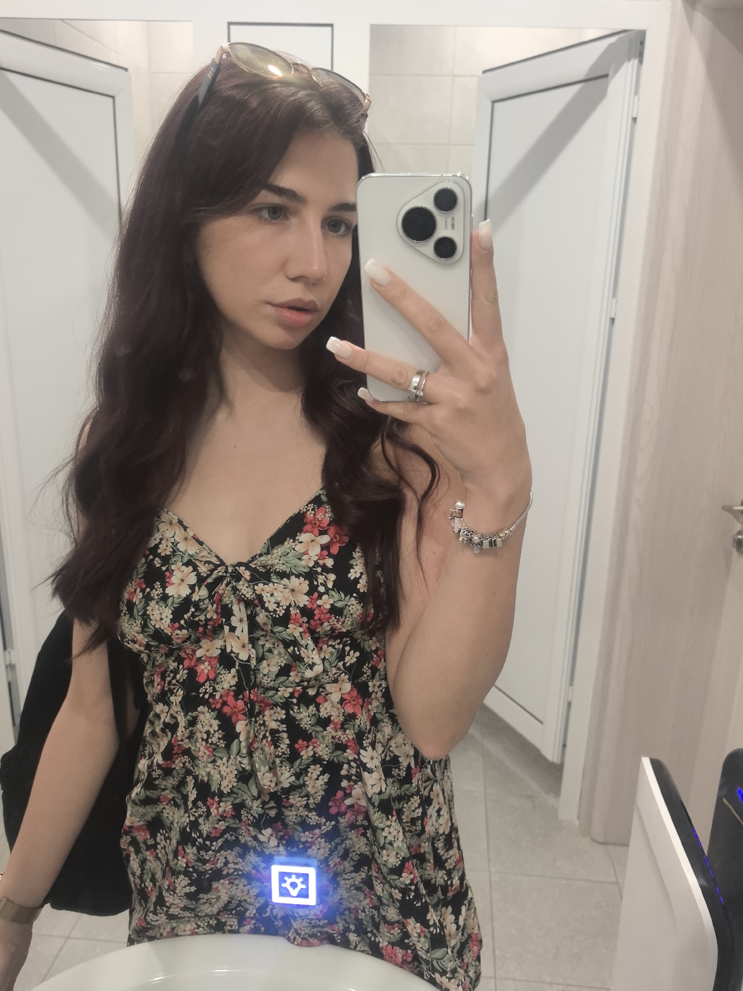 The image shows a young woman with long, dark, wavy hair taking a selfie in what appears to be a public restroom. She is wearing a floral print dress and sunglasses pushed up on her head. She has long, white acrylic nails and is wearing jewelry on her right hand, including rings and a charm bracelet. The background includes white doors, tiled walls and floor, and a white sink in the foreground. There is a blue illuminated icon with a sun-like symbol on the front of the sink area, possibly related to an automatic faucet or hand dryer. The overall scene is candid and suggests the woman is in a casual setting.