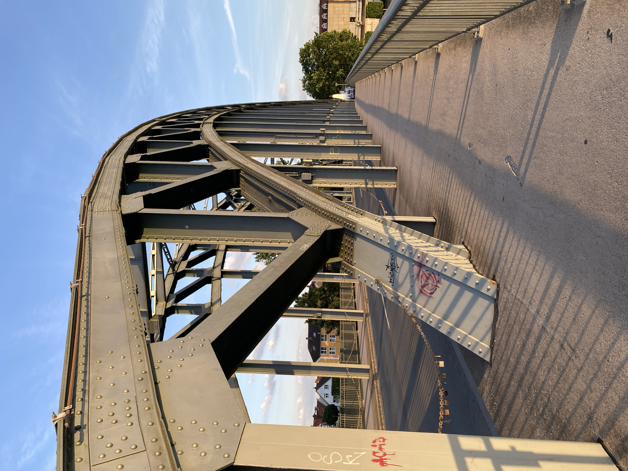 An image shows a close-up of a steel arch bridge over a road, with a pedestrian walkway on the right. The bridge is made of riveted steel beams forming an arch. The pedestrian walkway has a metal railing. Graffiti is visible on some of the bridge's support structures. In the background, there are buildings and trees under a blue sky with scattered clouds. A few padlocks are attached to a chain hanging from the bridge. This is the Weser-Rinteln Bridge in Rinteln, Germany.