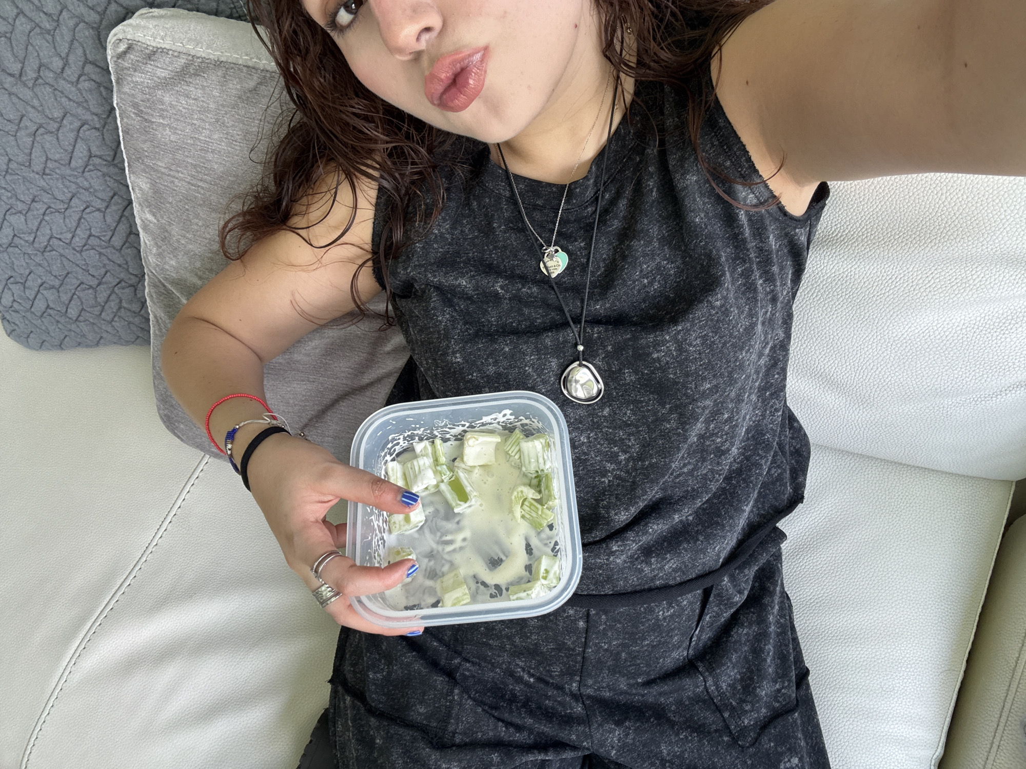 A woman is lying on a white leather sofa, holding a clear plastic container filled with what appears to be celery sticks in a creamy sauce. She is wearing a dark gray tank top and matching pants. She has brown curly hair and is wearing two necklaces. Her left wrist has several bracelets and rings adorn her fingers. She is looking at the camera with slightly pursed lips. A gray pillow rests behind her head. The image was taken in Santa Clara Ocoyucan, Mexico.