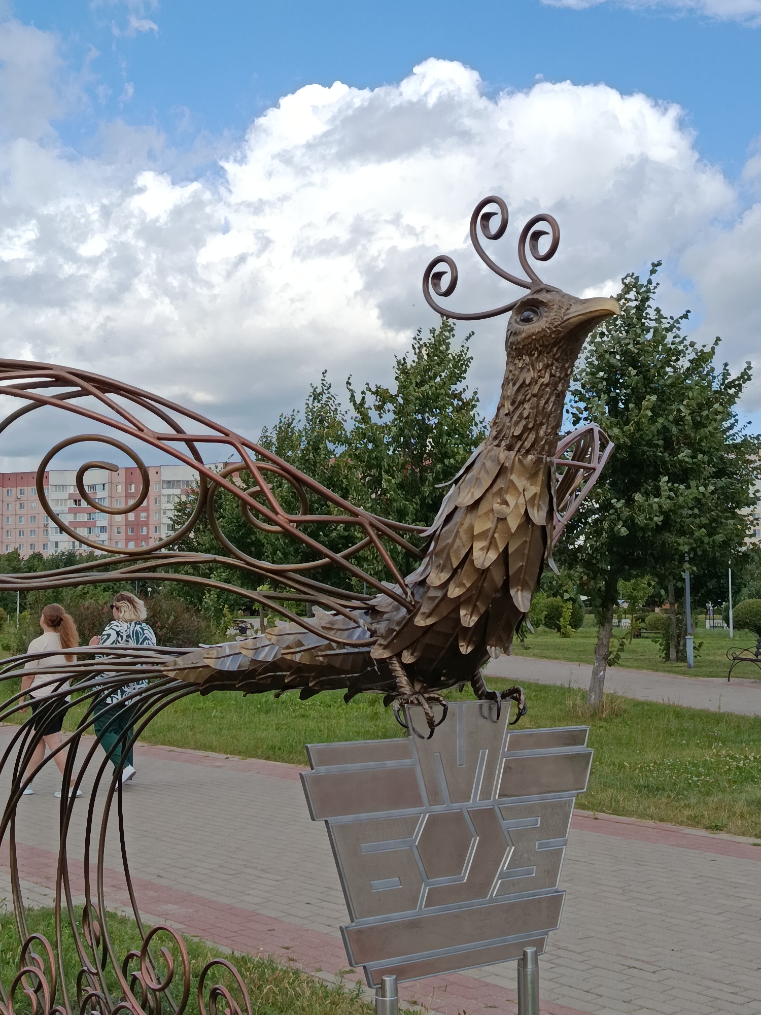 The image captures a metal sculpture of a bird, possibly a peacock or phoenix, situated outdoors in a park or public square. The sculpture is intricate, with detailed feather-like metal plates forming its body and a decorative, spiraling crest on its head. It stands on a metallic base with geometric engravings. In the background, a paved walkway with a red brick border is visible, leading towards a green lawn and trees. Two women are walking on the path in the middle ground. Further back, a multi-story residential building with a pale pink facade and numerous windows is visible. The sky above is partly cloudy with blue patches. The scene appears to be from the city of Zhlobin, Belarus, as suggested by the context.