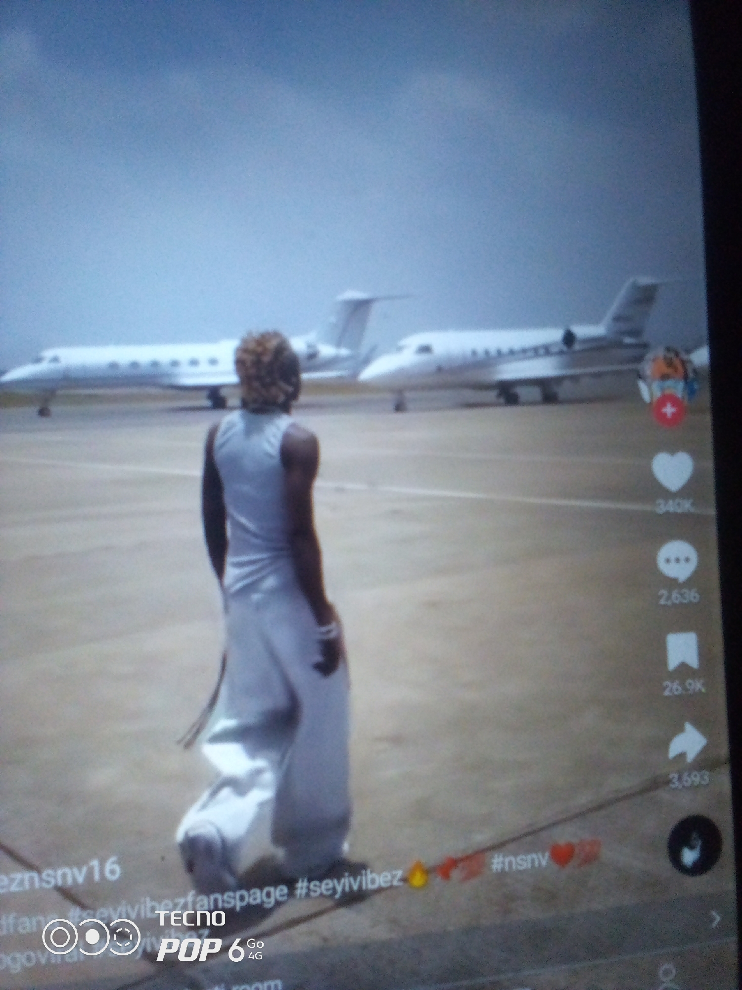 The image shows a person walking on an airport tarmac towards two private jets. The person is wearing a white outfit that drapes to the ground. The jets are white and appear to be parked side-by-side. The sky is overcast. The scene is likely from an airport in Howayi, Nigeria. The bottom of the image contains watermarks from social media platforms, including "TECNO POP 6 Go 4G".