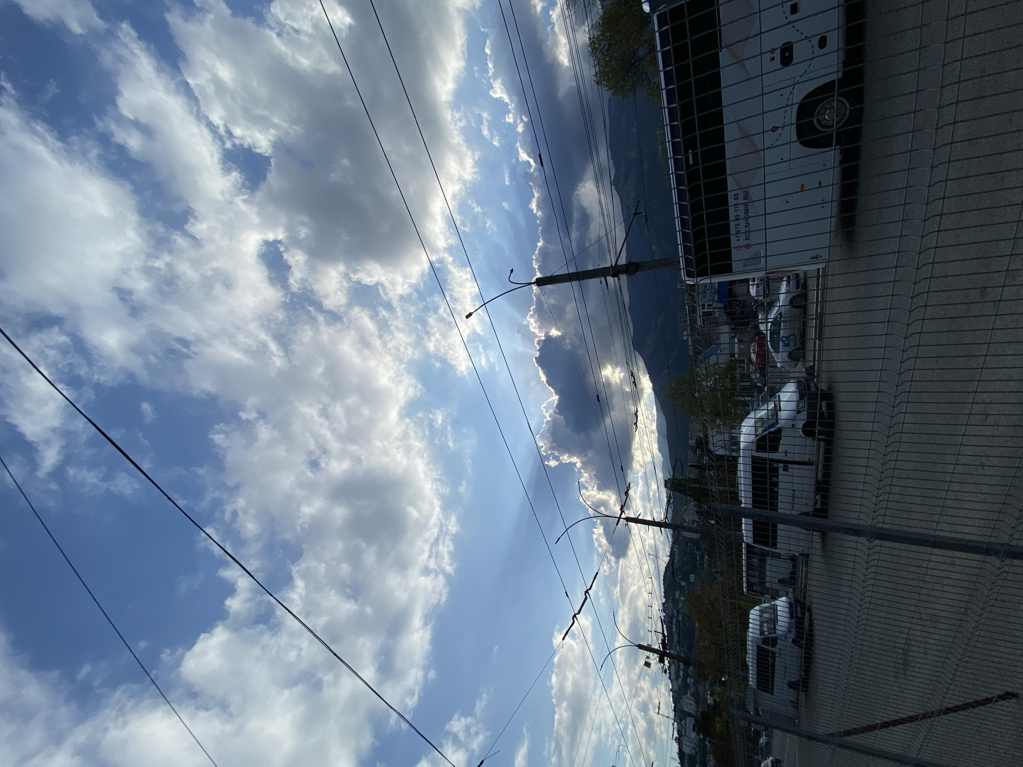 The image presents a view in Ялта, Ukraine, showing a fenced parking lot adjacent to a city street with power lines overhead. The sky, filled with cumulus clouds, dominates the left portion of the frame. Beyond the parking lot is a landscape of hills or mountains. A bus and several white vans are parked in the lot. The presence of power lines and overhead wiring suggests urban infrastructure.