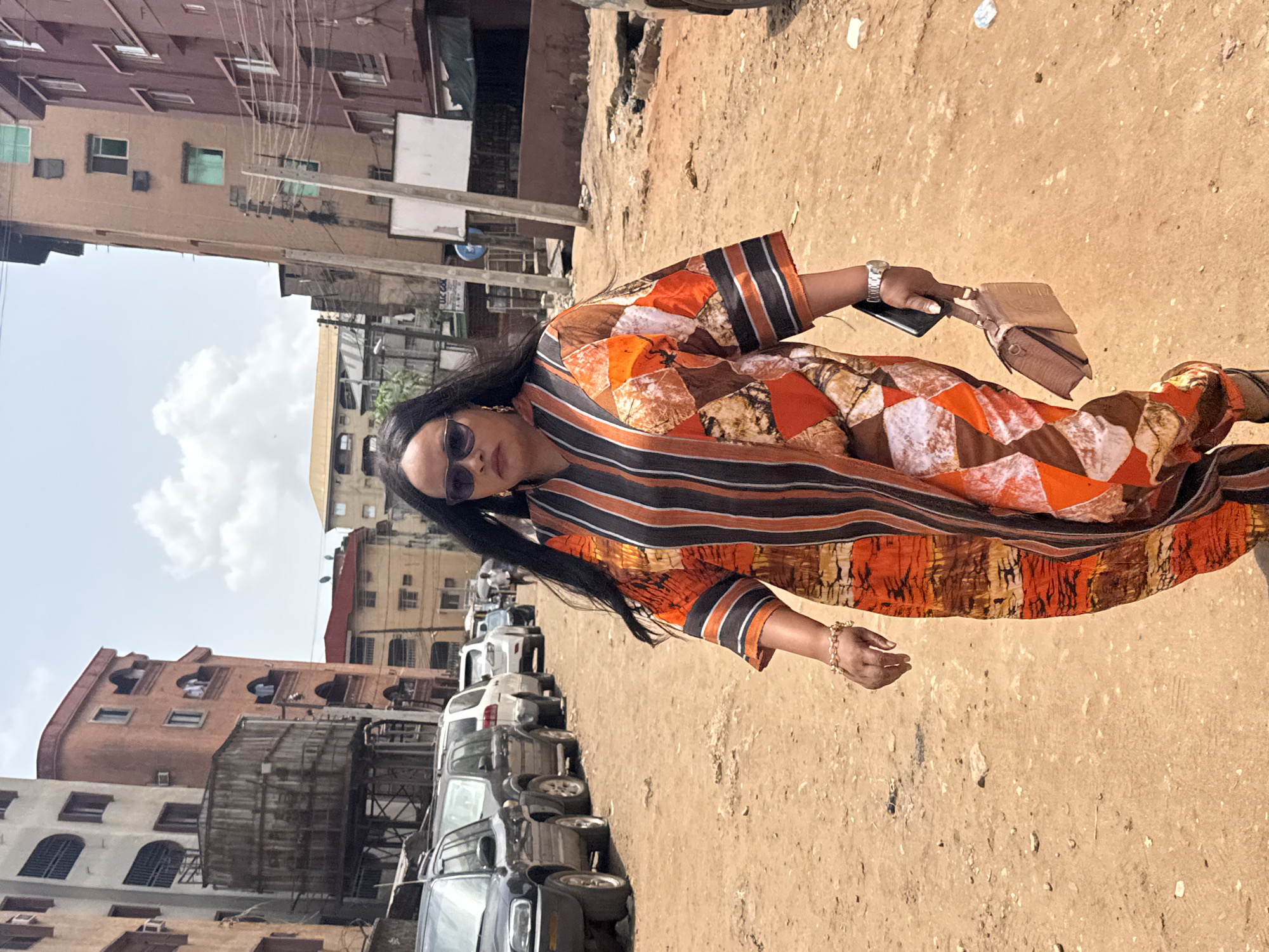 A woman stands on a dirt road lined with parked cars in Onitsha, Nigeria. She wears sunglasses, a watch, bracelets, and a long, colorful patterned dress. In her hand, she holds a phone and a clutch purse. Buildings of various heights and architectural styles are visible in the background, along with utility poles and wires. The sky is partly cloudy.