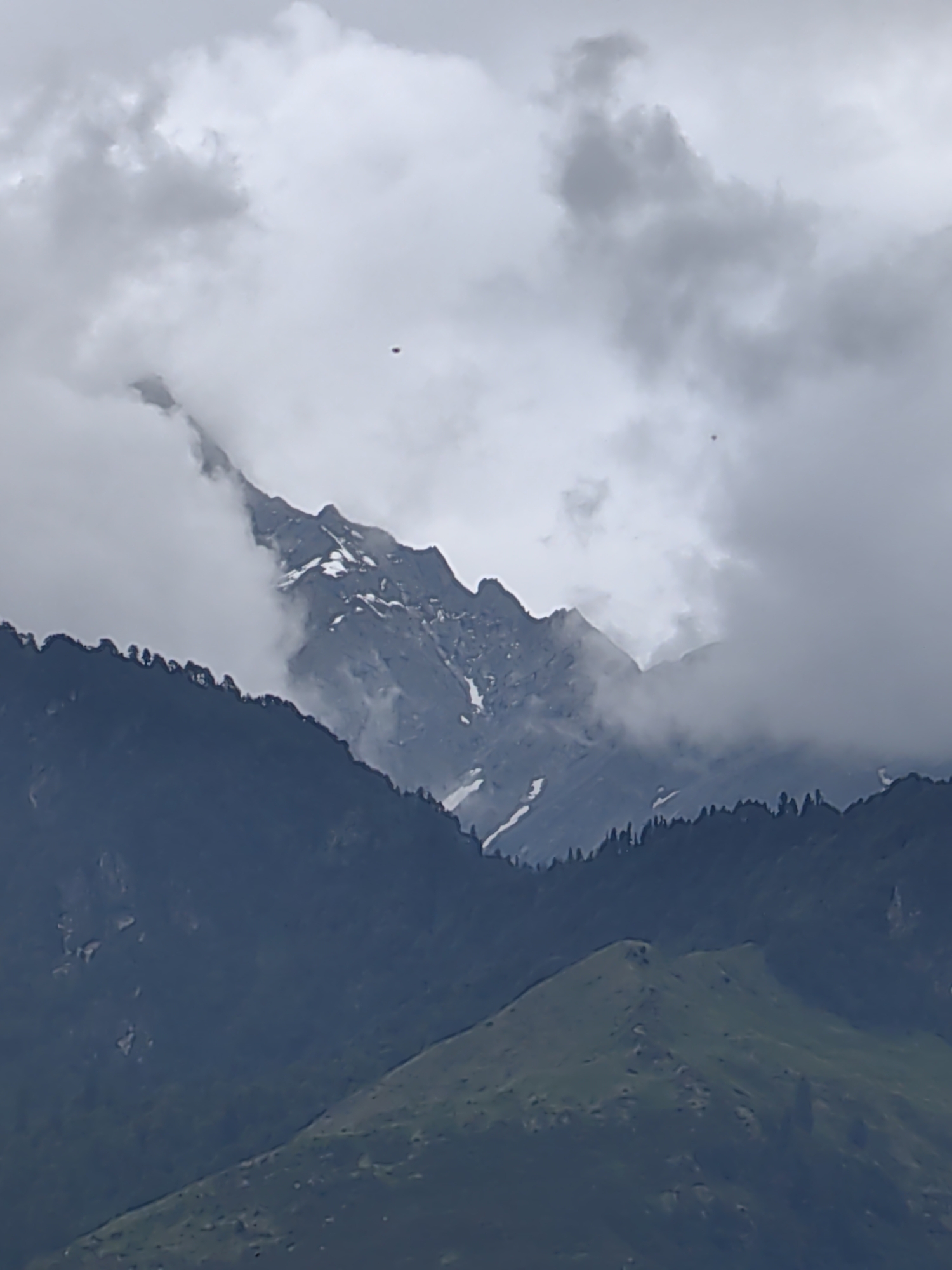 The image depicts a mountain landscape with dramatic cloud cover. In the distance, rugged, snow-capped peaks rise sharply, partially obscured by thick, grey clouds. The slopes of the lower mountains are covered in dark green foliage, with a line of coniferous trees visible along the ridgeline. In the foreground, a grassy, rounded hill slopes upwards, its surface dotted with small shadows or depressions. Two small, dark, indistinct objects are visible in the sky, one higher and to the left, the other lower and to the right. The overall scene suggests a cool, possibly overcast day in a mountainous region. The location is Vashisht, India.
