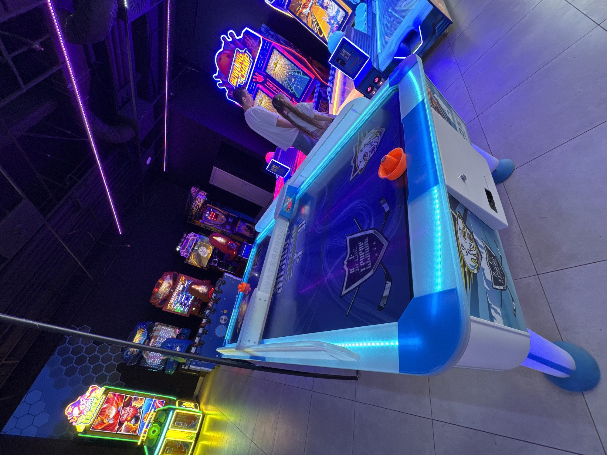 The image depicts an indoor amusement arcade or game center. Brightly colored arcade games are lined up against a wall, and an air hockey table with blue LED lighting is prominently featured in the foreground. Two people, a male and a female, are positioned near a bowling game, seemingly engaged in playing or interacting with it. The environment is lit with neon and LED lights, creating a vibrant and dynamic atmosphere typical of such entertainment venues. The location is not specifically indicated in the image, but it is stated that it is in the city of Podgorica, Montenegro. The tiled floor, arcade machines, and lighting suggest a commercial, recreational setting.