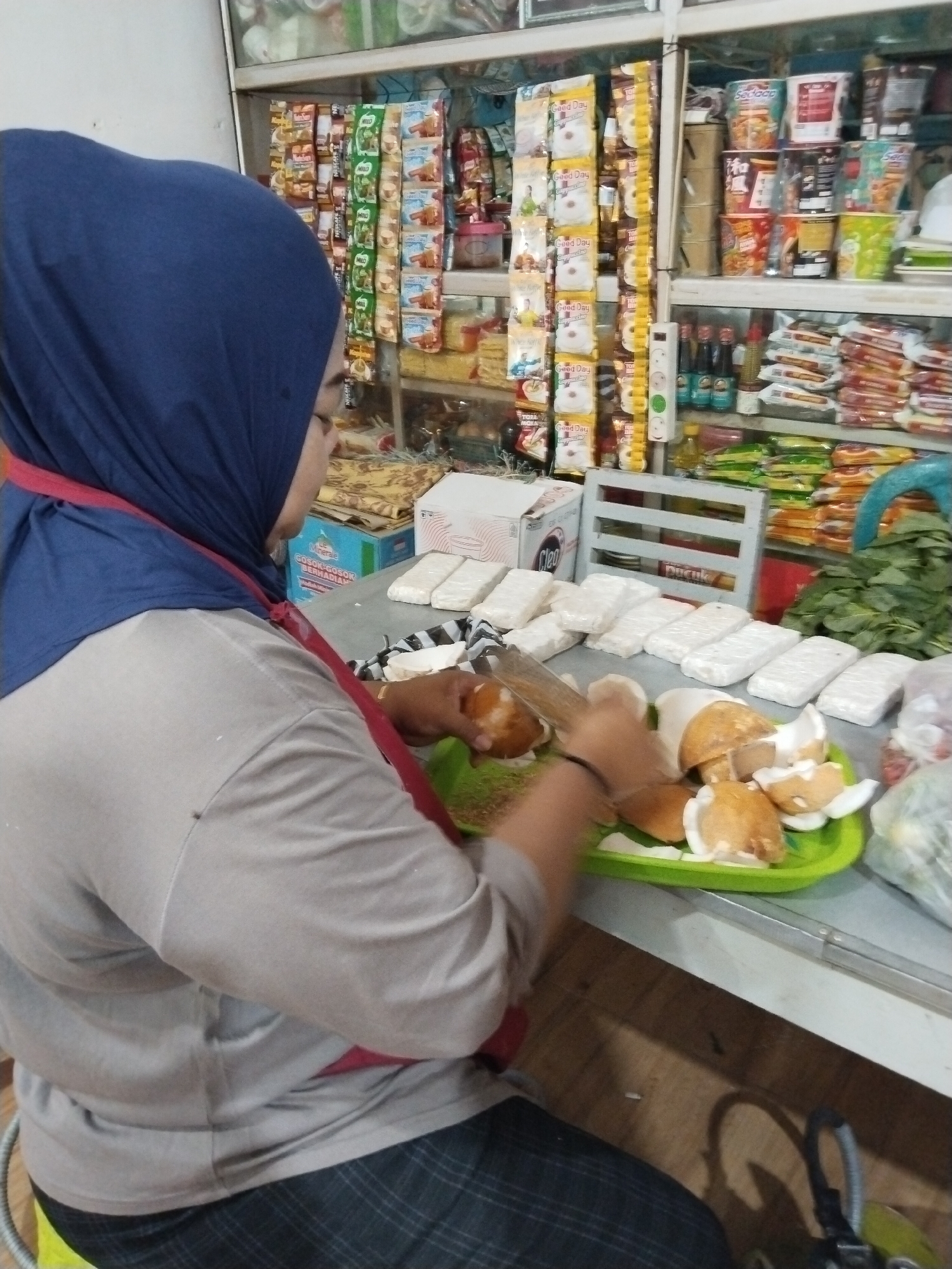 A woman, wearing a blue hijab and a grey t-shirt with a red accent, is seen from the side preparing coconuts at a market stall. She is using a wooden tool to split the coconuts, which are placed on a green tray. In front of her, on a grey counter, are several packages of white food items, possibly tempeh, and a box labeled "Cleo". Behind her, shelves are stocked with various packaged goods, including food items and drinks. The setting appears to be a small grocery store or market, likely in Prigi, Indonesia, given the context. The lighting is artificial, typical of an indoor environment.