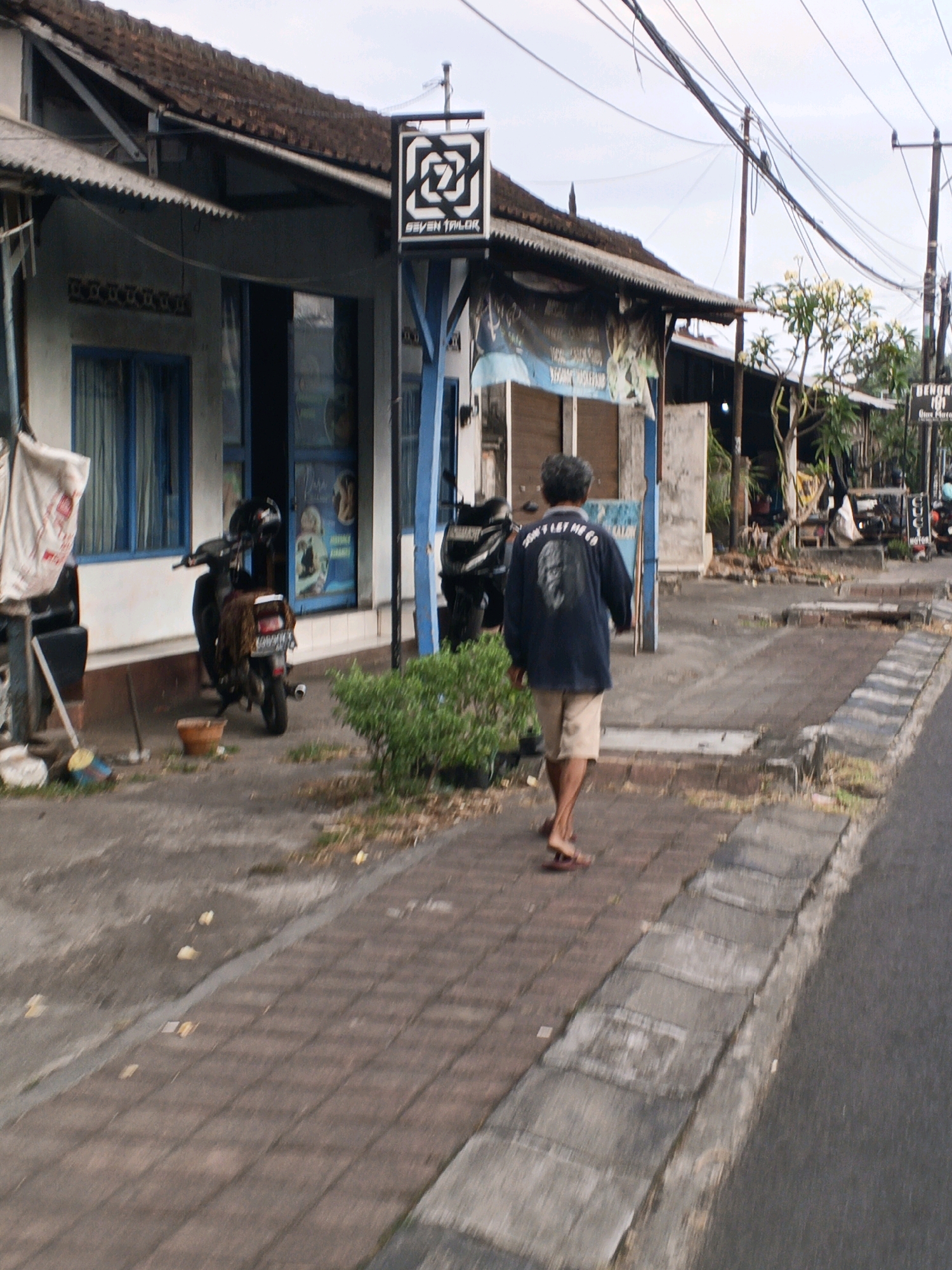 The image captures a street scene in Sukawati, Indonesia. A man wearing a dark blue long-sleeved shirt with text and a portrait on the back, and light brown shorts, walks away from the camera on a brick sidewalk. He is wearing dark flip-flops. To his left, a motorcycle is parked, covered with a patterned cloth. Further left, a building with a white facade and blue-framed windows is visible. A sign with a black and white geometric logo and the words "SEVEN TAILOR" hangs from a blue pole in front of the building. The background shows more buildings, trees, and utility poles with a tangle of wires overhead. The road runs alongside the sidewalk. The lighting suggests it is daytime, possibly overcast.