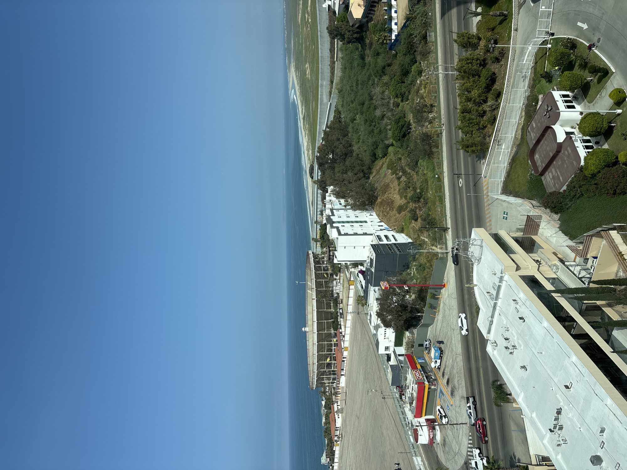 The image shows an aerial view in Tijuana, Mexico, including the coastline along the Pacific Ocean, buildings, a street, and vegetation. The ocean occupies a significant portion of the left side, meeting the beach and adjacent buildings. The buildings are various types, including residential and commercial structures, some of which are under construction. A street runs parallel to the coastline, featuring moving cars and trucks. Vegetation including trees, shrubs, and grass are also visible.