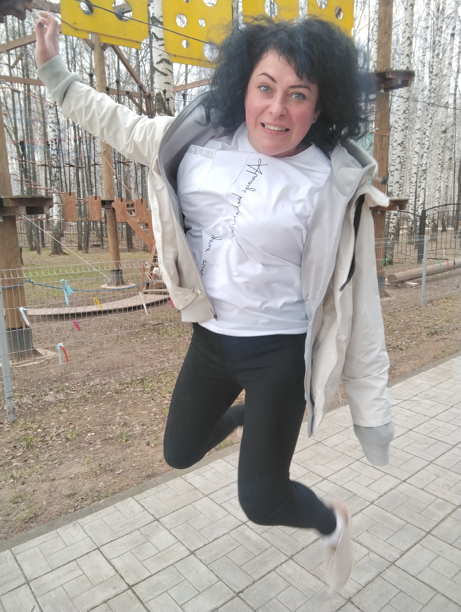 A woman is captured mid-air, jumping with an excited expression in an outdoor setting, likely an adventure park in Chaikovsky, Russia. She has dark, curly hair and blue eyes, with her mouth slightly open, showing teeth, conveying a sense of fun or surprise. She wears a white t-shirt with unreadable cursive text, possibly "forever your own", on the left side of her chest, a light grey or beige jacket worn open, and black leggings. Her arms are raised and spread as she leaps.
The background features numerous tall, slender birch trees, mostly bare, indicating late winter or early spring. Interspersed among the trees are elements of a ropes course, including wooden platforms, ropes, and distinct bright yellow obstacles shaped like cheese with holes. A low metal mesh fence, adorned with various colorful ribbons (blue, pink, red, yellow), runs horizontally across the mid-ground. The ground behind the fence is a mix of dirt, dried leaves, and sparse grass. In the foreground, a paved path made of light grey rectangular pavers provides the surface from which she has jumped. The lighting suggests an overcast or diffused daylight.