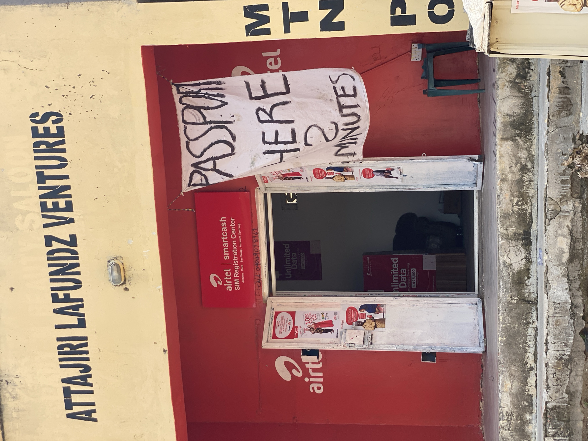 The image depicts the exterior of a business establishment in Jalingo, Nigeria, identified by the signage "ATYAJIRI LAFUNDZ VENTURES" and the presence of "Airtel" and "MTN" branding, indicating it is likely a telecommunications service provider or an agent. The facade is painted a pale yellow and red. Black stenciled letters spell out "ATYAJIRI LAFUNDZ VENTURES" across the top. Below this, a red awning extends over the entrance.
The entrance itself is divided into two sections, with the left side displaying the "airtel" logo. The right side of the entrance is open, revealing an interior with red walls and signs advertising "Unlimited Data." A white banner hangs prominently in front of the entrance, with hand-painted black text that reads "PASSPORT HERE 2 MINUTES." This banner suggests a service related to passport applications or processing is offered at this location.
Within the doorway, on the left side, there are two white doors, one slightly ajar. These doors have posters affixed to them. The posters appear to be advertisements for Airtel services, featuring images of people and text in what looks like Hausa or a similar Nigerian language. One poster mentions "ZERO CHARGE" and "10 BIRR," while another shows a woman in traditional attire and text about "KUDIR DA KA."
Inside the open doorway on the right, a dark interior is visible. There are signs within, one of which displays "Unlimited Data" and a price of "N25,000." A dark-colored armchair is partially visible in the background.
To the right of the entrance, on the exterior yellow wall, large black letters spell out "MTN." Further to the right, a small green plastic stool is placed near the wall, and an electrical outlet is visible on the red wall.
The ground in front of the establishment consists of rough, weathered concrete steps leading down to a slightly uneven surface. Some debris and dry vegetation are present on the ground and steps. The overall impression is of a busy, functional commercial space in an urban environment, likely catering to common mobile network and potentially passport-related services.