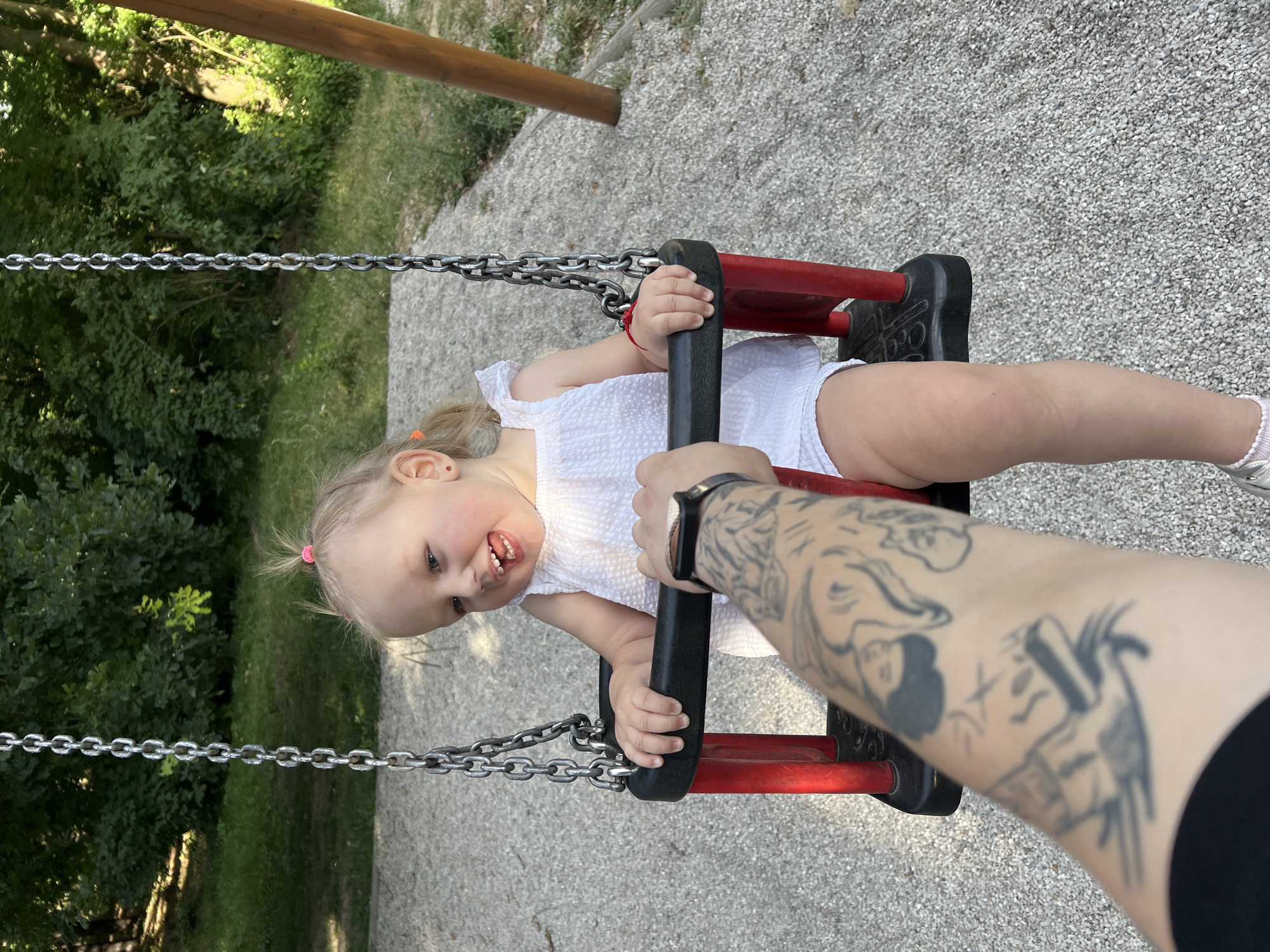 A young Caucasian girl, approximately 2-3 years old, with blonde hair in two small pigtails, is seated in a red and black swing at a playground. She is wearing a white, textured romper and has red earrings and a red string bracelet on her left wrist. Her mouth is open, and she appears to be smiling or laughing. Her right hand is resting on the swing's safety bar, and her left hand is near her face. A tattooed arm, presumably belonging to an adult, is visible in the foreground, reaching towards the girl and holding what appears to be a smartwatch. The background consists of a gravel-covered ground, trees, and foliage, indicative of an outdoor park setting. The image is captured in Pilsen, Czechia.
